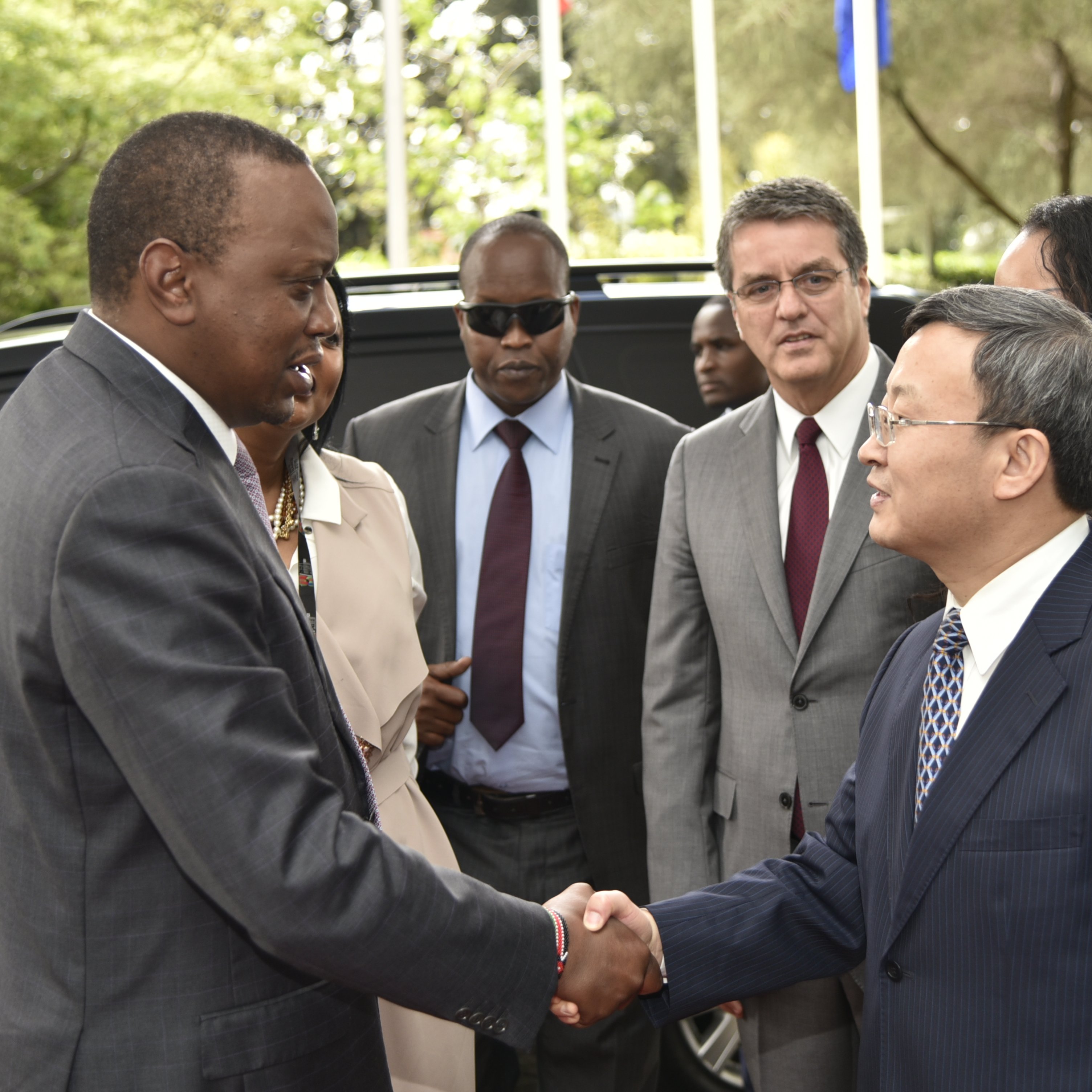 L'image montre une scène de rencontre officielle à l'extérieur, dans un environnement verdoyant avec des arbres et des drapeaux en arrière-plan. Au centre, deux hommes se serrent la main. L'un est habillé en costume sombre, avec une expression sérieuse, tandis que l'autre porte une cravate et un costume clair. En arrière-plan, d'autres personnes observent la rencontre, certaines avec des lunettes de soleil, indiquant un moment formel. L'atmosphère est celle d'une discussion diplomatique.