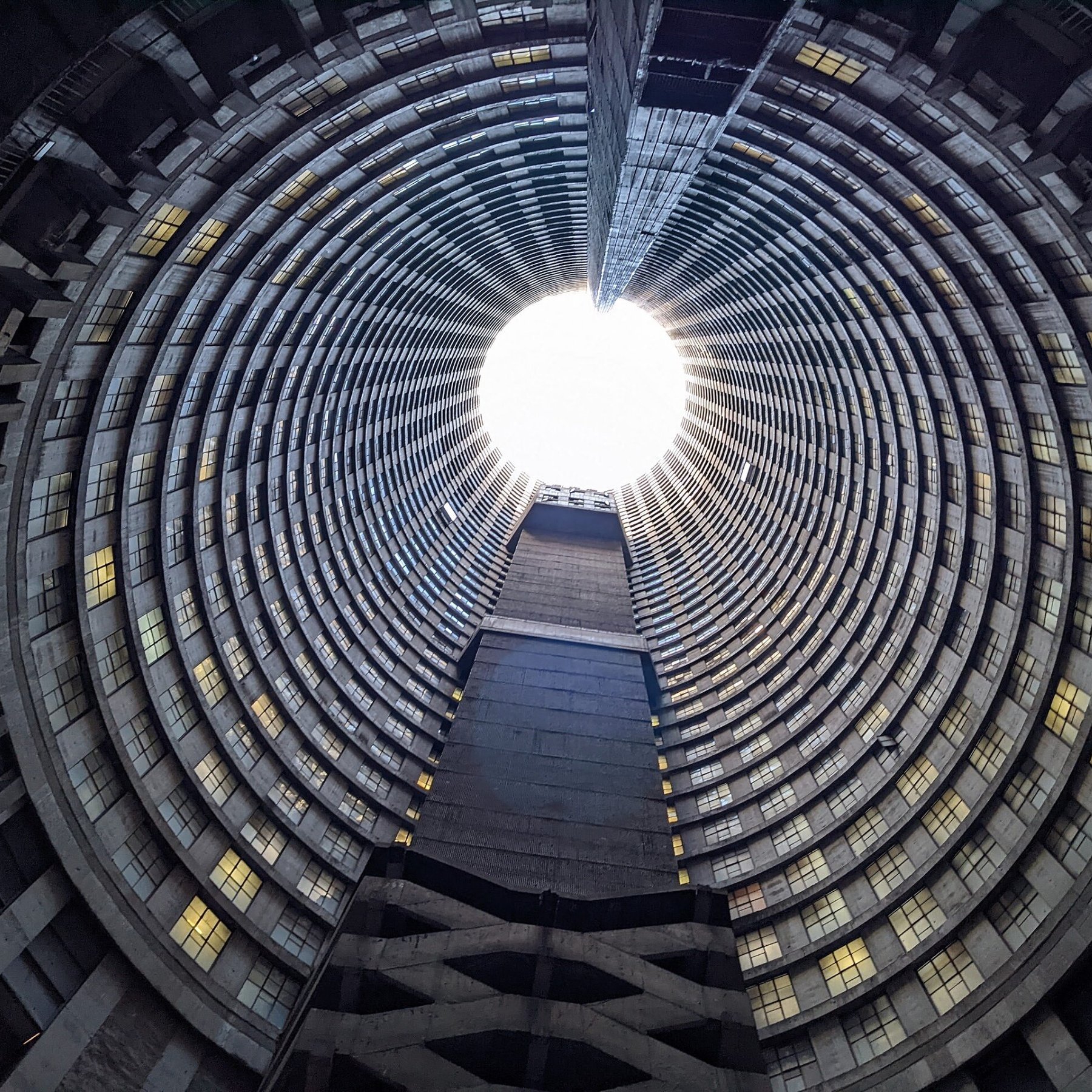 Imaginez vous tenir au centre d'une immense structure circulaire. La vue se dirige vers le haut, où un puits de lumière éclaire l'espace depuis l'ouverture au sommet. Les parois de ce bâtiment sont composées de béton et forment des motifs en spirale, créant une impression de profondeur et de hauteur. Les fenêtres qui bordent les étages, bien que nombreuses, laissent passer une lumière diffuse qui ajoute une sensation d'apaisement. Le sol est plat et peut sembler rugueux, contrastant avec la douceur de la lumière qui découpe le vide en haut. L'atmosphère est à la fois vide et pleine de possibilité, un mélange d'architecture industrielle et de nature, le tout se rejoignant dans ce grand espace ouvert.