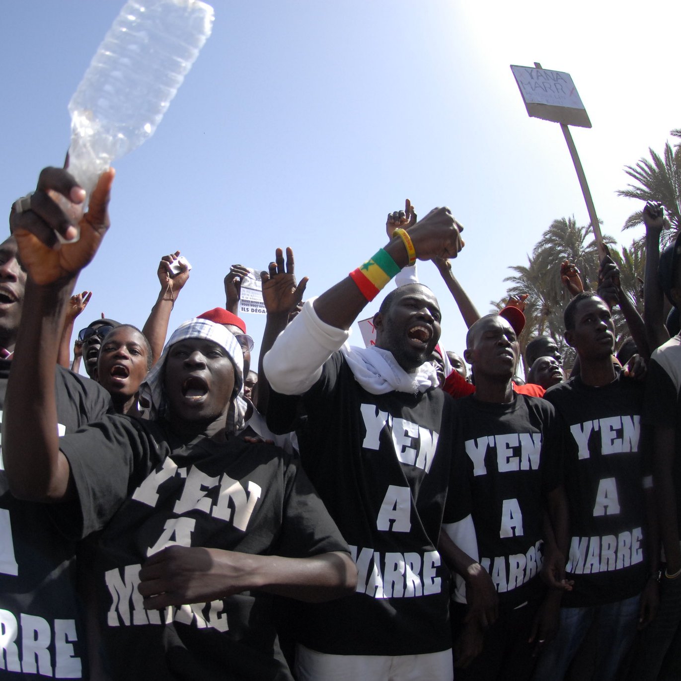 L'image montre un groupe de personnes rassemblées pour exprimer leur mécontentement, probablement lors d'une manifestation. Les membres du groupe portent des t-shirts noirs avec les mots "Y'en a marre" inscrits en lettres blanches, ce qui signifie qu'ils en ont assez d'une situation particulière. On peut les voir lever les bras, certains brandissant des bouteilles en plastique, et afficher des visages déterminés. En arrière-plan, on aperçoit des palmiers et un ciel bleu, indiquant une ambiance ensoleillée. L'énergie de la scène est palpable, avec des cris et une forte intensité émotionnelle.