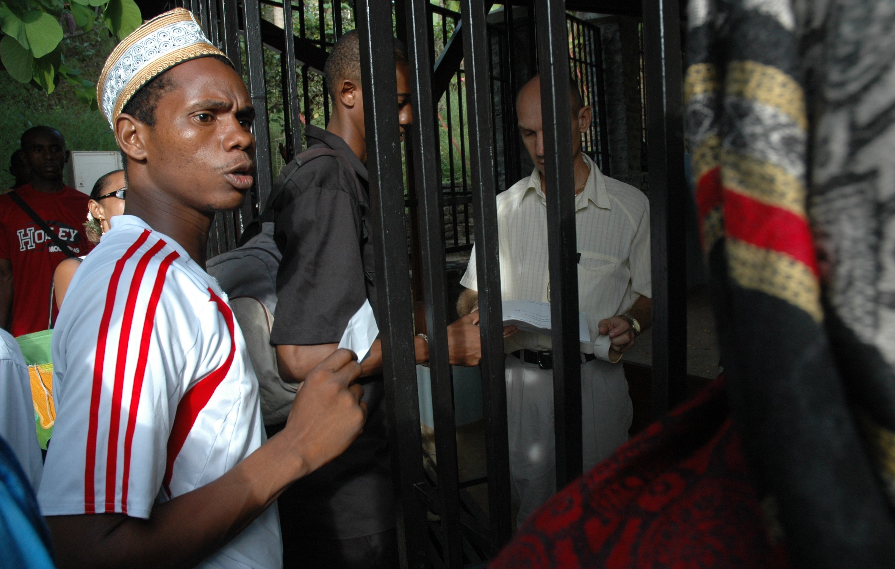 L'image montre un groupe de personnes rassemblées autour d'une structure métallique, probablement une grille ou une clôture. Un jeune homme au premier plan, portant un maillot blanc avec des rayures rouges et un chapeau traditionnel, exprime une certaine agitation ou curiosité. À côté de lui, d'autres personnes, dont un homme en chemise claire, semblent discuter ou échanger des documents. L'environnement est extérieur et verdoyant, suggérant un lieu fréquenté. Les expressions des visages indiquent une interaction dynamique et engageante entre les personnes présentes.