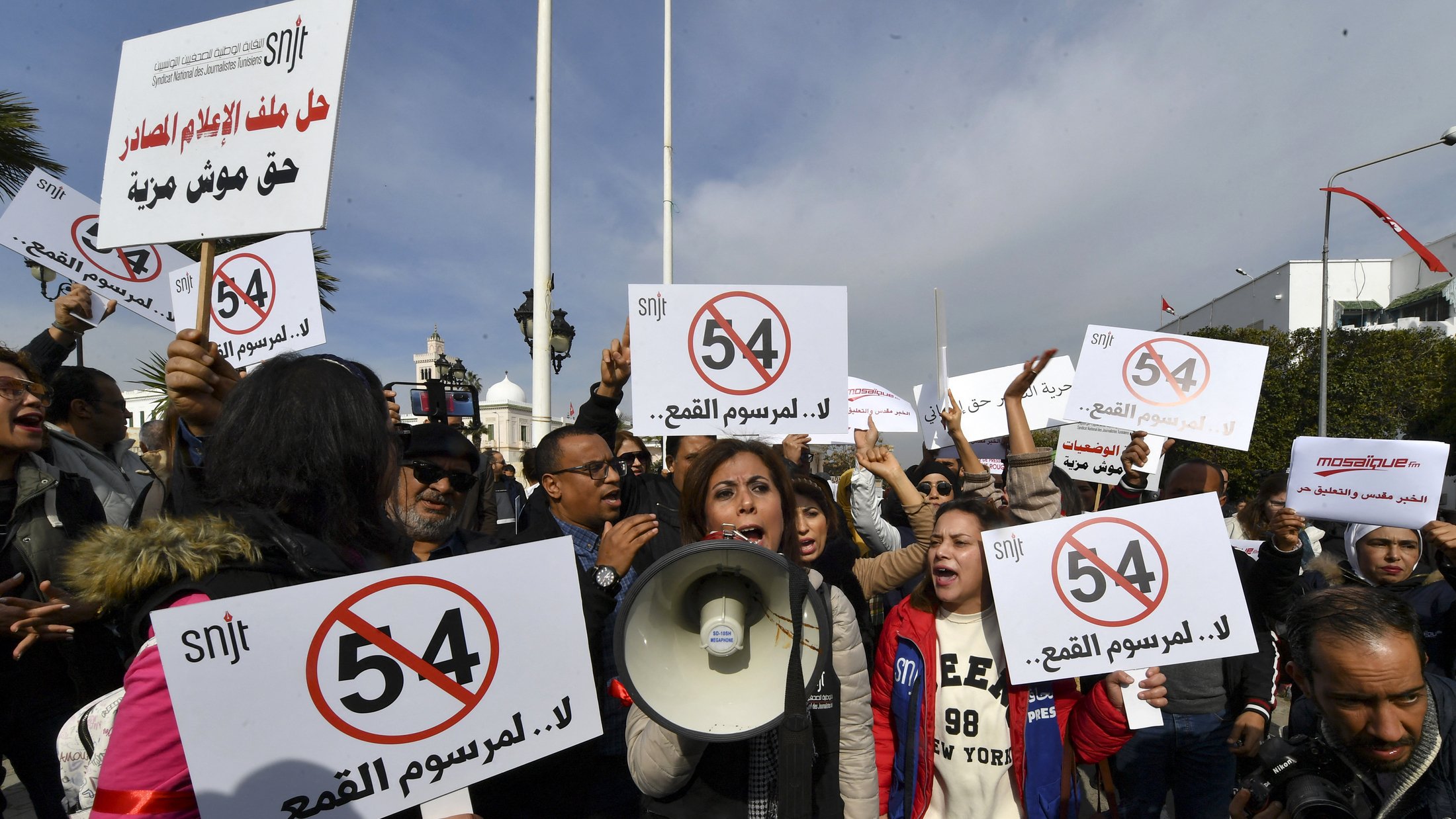 The image depicts a protest scene with a group of people holding signs and banners. In the foreground, a woman is speaking into a megaphone, likely leading the demonstration. The signs feature the number "54" and messages opposing censorship, suggesting that the protest is focused on issues related to freedom of expression or press freedom. The crowd appears to be actively engaged in the protest, raising their signs and voicing their demands. The setting seems to be outdoors, with flags and possibly a government building in the background.