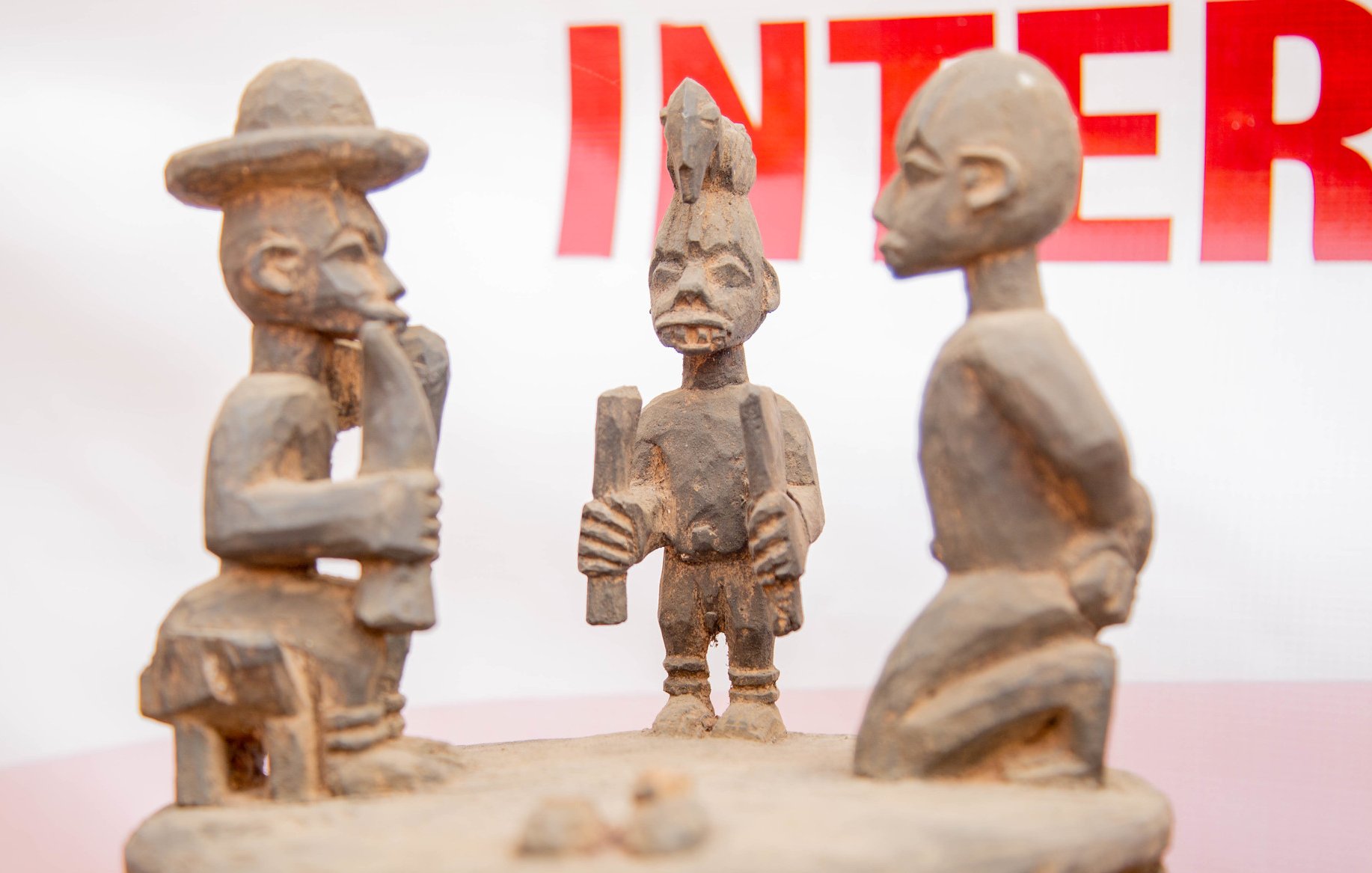 L'image présente une sculpture en bois représentant trois figures humaines stylisées. Au centre, une figure masculine, aux traits marqués, tient un objet dans les mains, probablement un outil ou un symbole culturel. Il est entouré de deux autres sculptures, l'une à gauche, qui porte un chapeau large, et l'autre à droite, avec une coiffure distinctive. Les personnages semblent engagés dans une conversation ou un échange, ce qui suggère une interaction sociale. Les détails des sculptures, comme les expressions des visages et les postures, témoignent d'un art tribal riche en histoire et en culture. Le fond de l'image est flou, mettant l'accent sur les sculptures elles-mêmes.