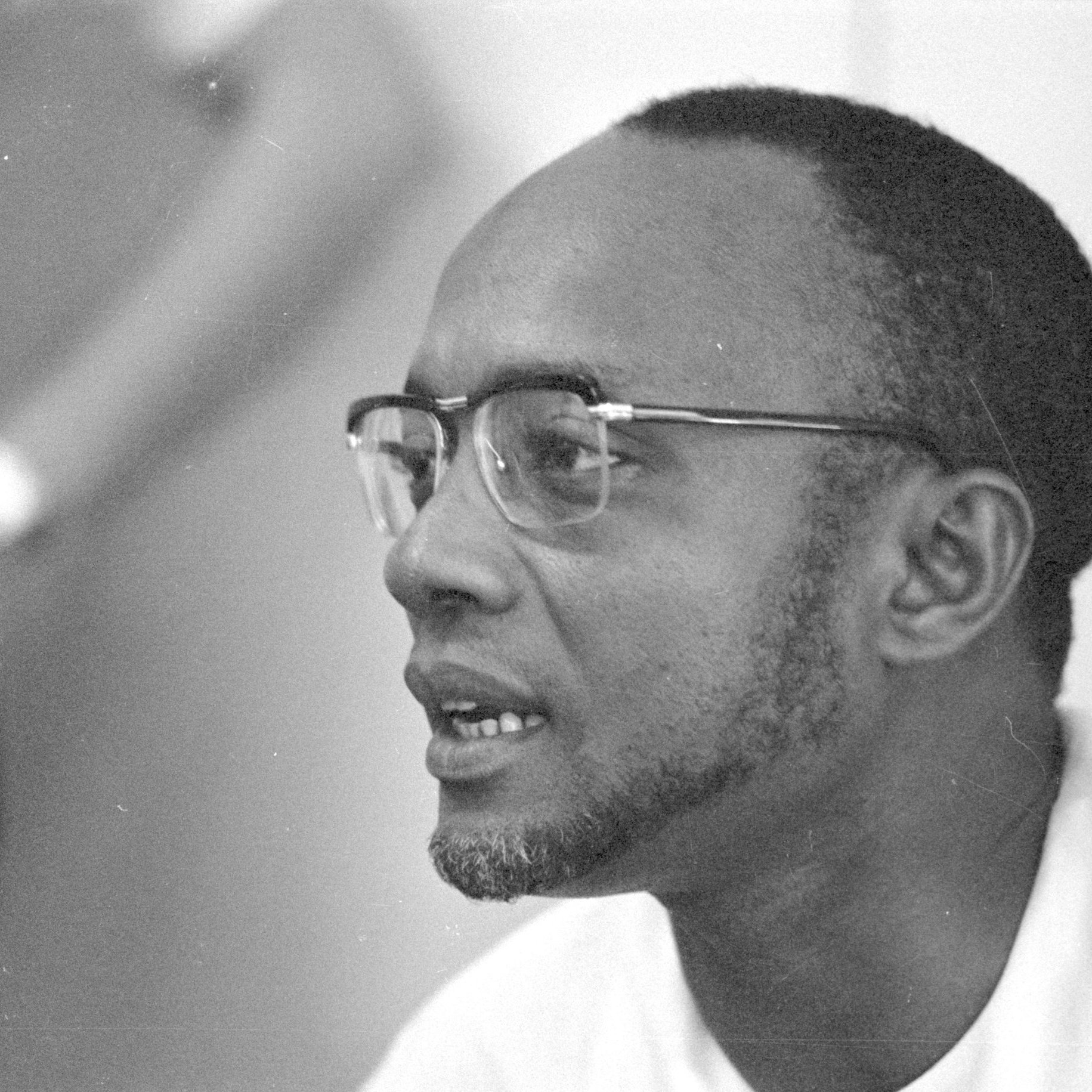 L'image représente un homme portant des lunettes à monture fine. Il a un visage allongé avec une petite barbe bien taillée. Ses cheveux sont courts et légèrement bouclés. L'homme semble concentré et engagé dans une conversation. En arrière-plan, on peut distinguer une silhouette floue d'une autre personne, suggérant qu'il se trouve dans un environnement social ou professionnel. L'éclairage est doux, ce qui met en valeur les traits de son visage.