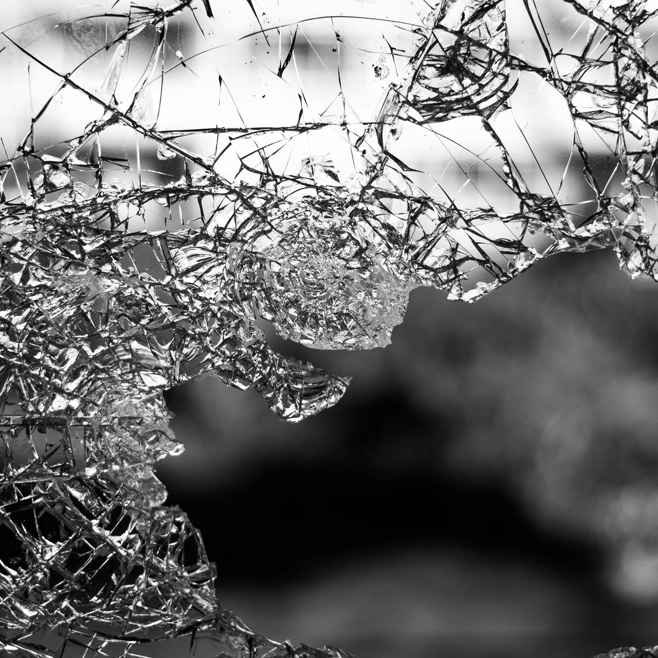 L'image montre un morceau de verre brisé, dont les nombreux éclats forment des motifs irréguliers. On peut ressentir la fragilité et la netteté des bords du verre. La lumière semble traverser certains éclats, créant des jeux d'ombre et de lumière. L'arrière-plan est flou, évoquant une ambiance sombre et mystérieuse. Cette composition souligne la beauté inattendue du verre cassé, tout en évoquant une sensation de vulnérabilité.