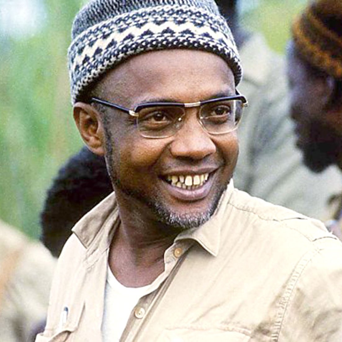 L'image montre un homme souriant, avec des lunettes au cadre fin et des cheveux rasés, portant une chemise beige. Il a une barbe grisonnante et son expression est chaleureuse et confiante. Il porte un bonnet tricoté, typique, dont les motifs évoquent un style traditionnel. En arrière-plan, plusieurs silhouettes floues d'autres hommes, habillés de manière similaire, suggèrent une ambiance de camaraderie et de travail en équipe. La scène véhicule une sensation d'optimisme et de détermination.