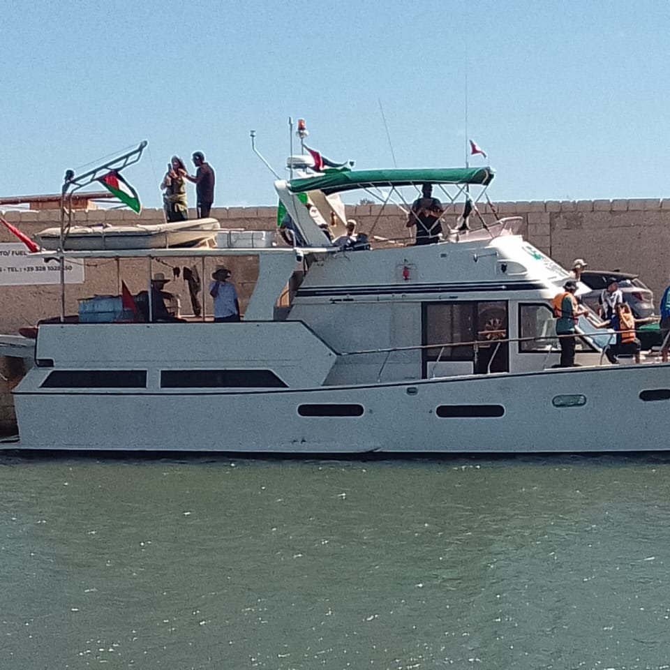 The image shows a large motorboat docked at a marina. The boat has a white hull with a green and white canopy on top. A group of people can be seen on the boat and along the dock, some engaging in conversation and others preparing to board. The surrounding area appears sunny, and there are flags displayed on the boat and nearby structures. The water is calm, reflecting the bright blue sky.