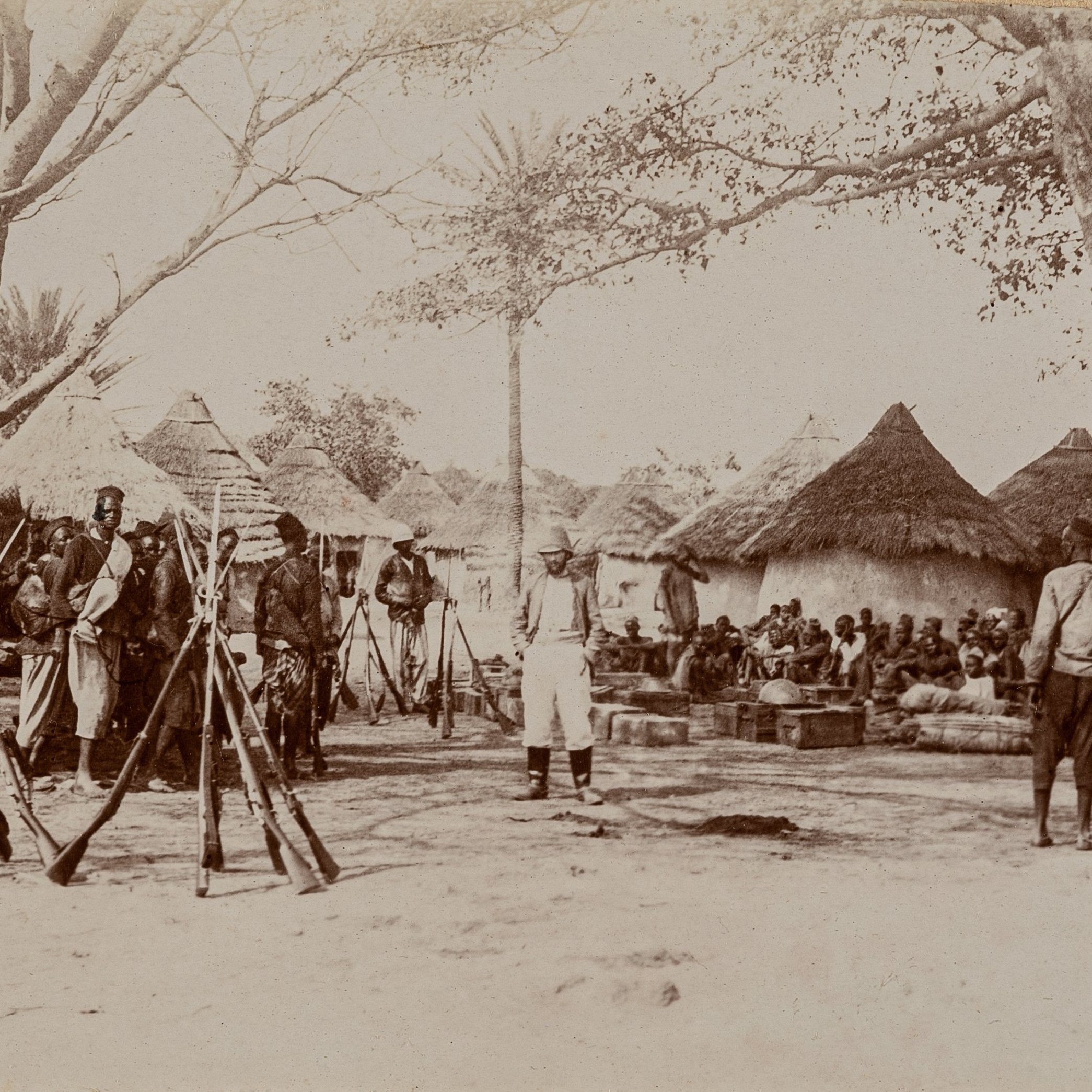 L'image représente une scène historique à l'extérieur d'un village. Au premier plan, plusieurs fusils sont posés verticalement sur le sol, formant une sorte de cadre. Des hommes, en majorité vêtus de vêtements traditionnels, sont rassemblés autour d'une zone centrale. Certains semblent discuter tandis que d'autres restent attentifs, leur posture indiquant un moment de rassemblement communautaire ou de célébration. En arrière-plan, on aperçoit des huttes de forme circulaire, couvertes de toits en paille, typiques des constructions de certaines régions rurales. Plusieurs arbres offrent de l'ombre, ajoutant à l'ambiance naturelle de l'endroit. Le sol est poussiéreux, indiquant le climat aride de la région. Les expressions des visages reflètent un mélange d'intérêt et de sérieux, suggérant une rencontre importante au sein de la communauté.