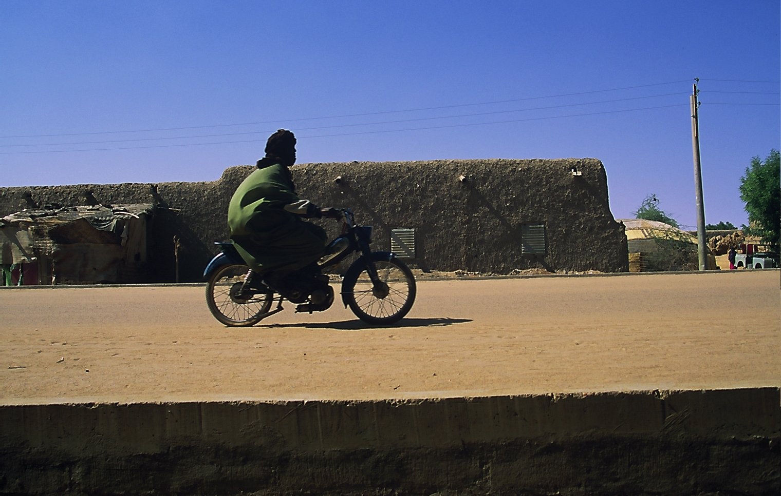 L'image montre un homme en train de rouler à bicyclette sur une route en terre. Il porte une tenue traditionnelle de couleur verte, qui contraste avec le décor. En arrière-plan, on peut apercevoir des bâtiments en adobe, typiques des régions désertiques, avec des murs de terre et des fenêtres. Le ciel est d'un bleu éclatant, sans nuages, et l'atmosphère générale est calme, suggérant une journée ensoleillée. La lumière du soleil éclaire la scène, mettant en valeur les textures du sol et des constructions.
