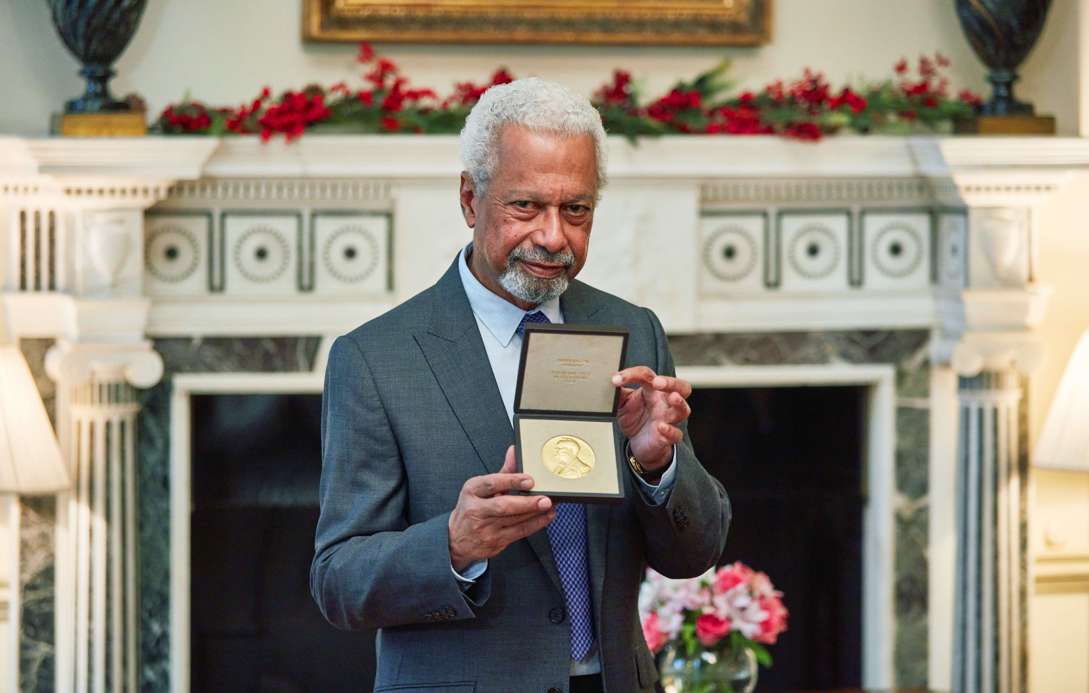 L'image montre un homme âgé, avec des cheveux poivre et sel et une barbe, qui porte un costume gris. Il se tient fièrement devant une cheminée décorée, tenant une médaille en or dans un écrin, qu'il montre à la caméra. En arrière-plan, on peut distinguer une décoration florale avec des fleurs disposées sur une table, créant une ambiance chaleureuse et élégante. La pièce est bien éclairée et a un air prestigieux, suggérant une occasion spéciale.