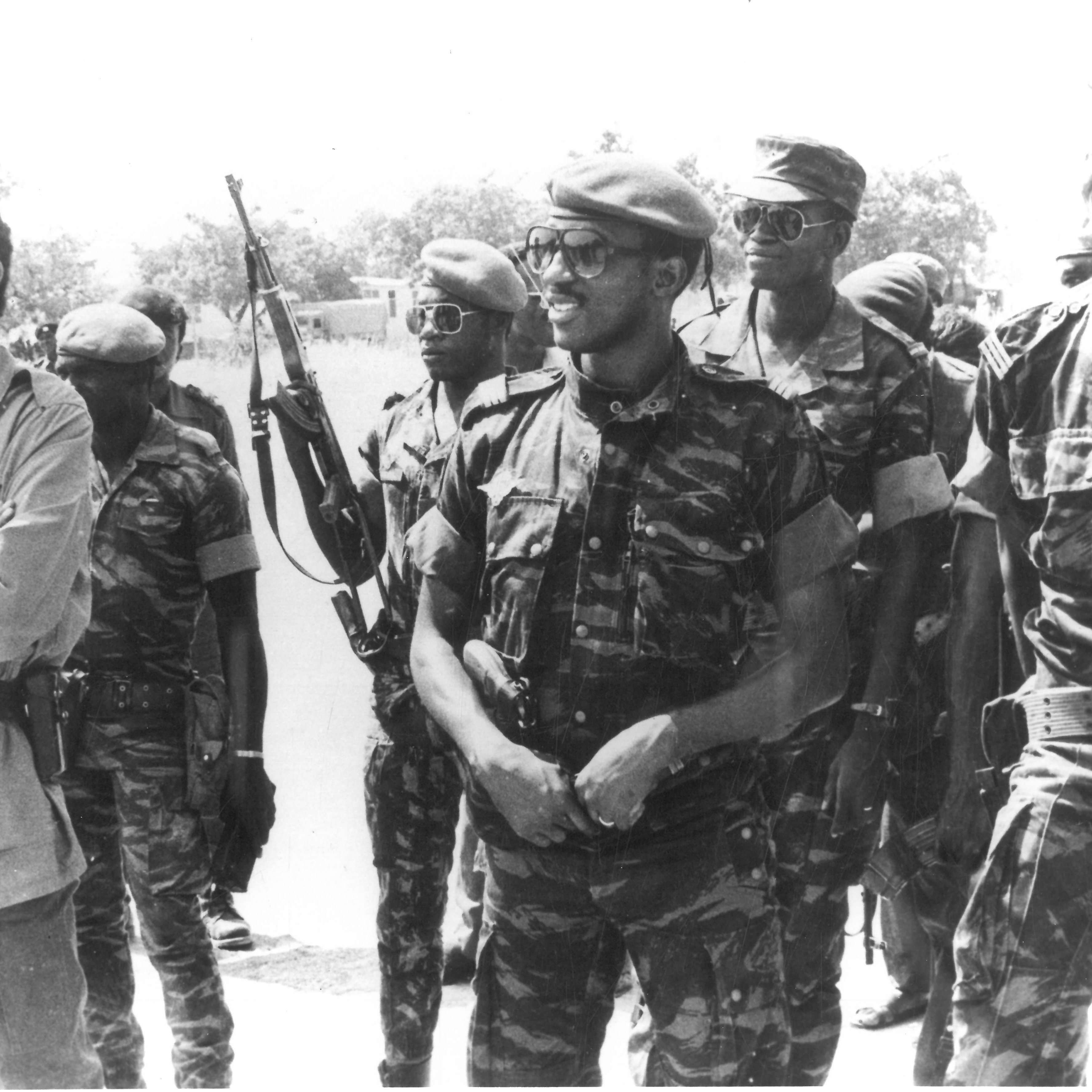Cette image en noir et blanc montre un groupe de soldats, tous habillés en uniforme militaire. Au centre, un homme souriant porte un beret et un uniforme camouflage. À sa droite, un autre homme, également en uniforme, affiche un large sourire. En arrière-plan, plusieurs soldats tiennent des fusils et portent des lunettes de soleil. L'ambiance semble détendue, avec des expressions de camaraderie parmi les membres du groupe. Le décor en arrière-plan est peu détaillé, mais on peut deviner un environnement extérieur.