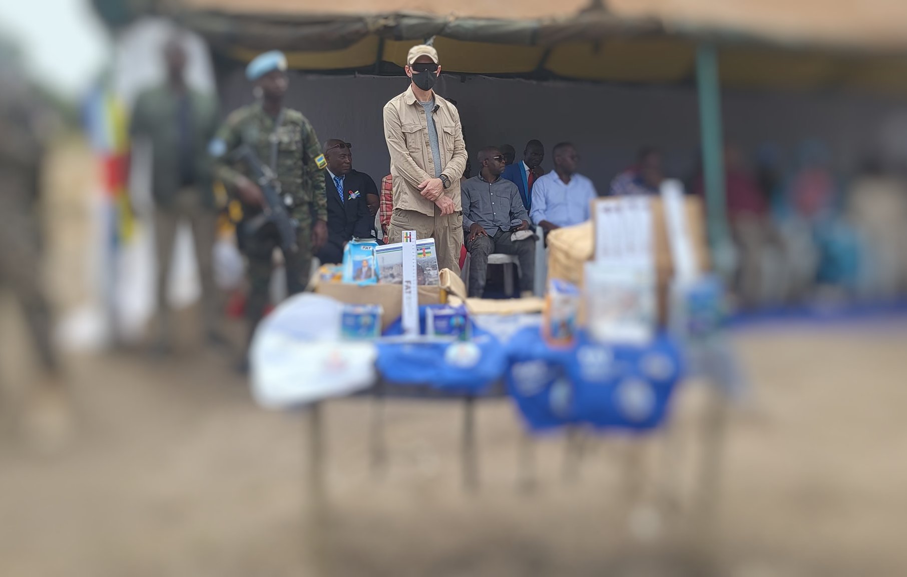 L'image montre un événement en plein air, avec une tente sur le côté. Au centre, on voit une table couverte d'objets et de brochures, probablement liés à une présentation ou un programme. À l'arrière-plan, plusieurs personnes sont assises, y compris des enfants et des adultes. Un homme se tient debout, portant une casquette et un masque, semblant être un intervenant ou une personne d'autorité. La scène reflète un moment de rassemblement communautaire, chargé d'une atmosphère d'échange et d'information.