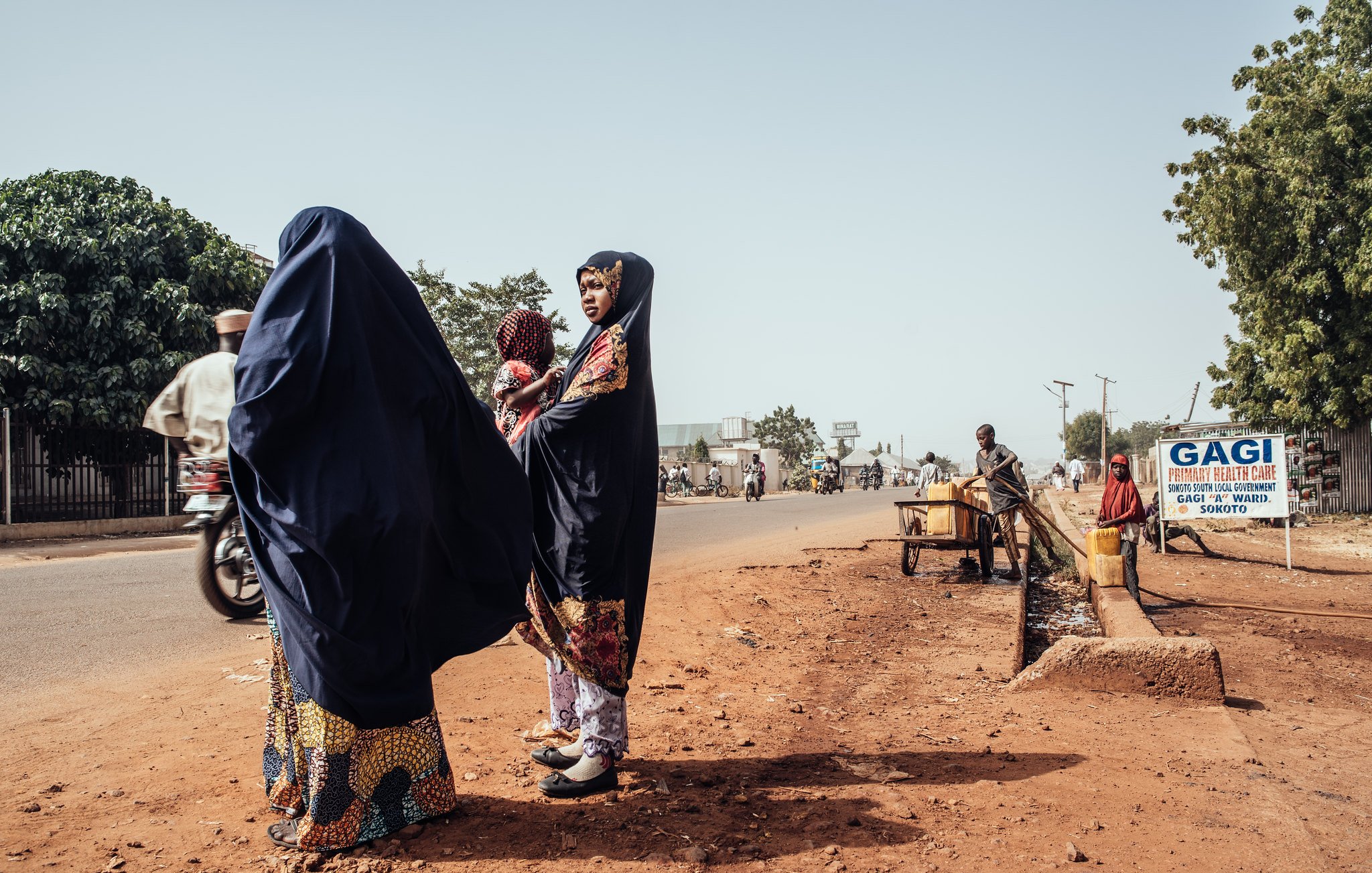 Dans cette image, nous sommes sur une route dans un environnement urbain. Sur le côté gauche, deux femmes portant des vêtements traditionnels de couleur sombre se tiennent ensemble. L'une d'elles a un sourire et semble discuter. Elles sont entourées de terre brune, et l'atmosphère est calme. Sur le côté droit, un homme est assis près d'un petit chariot, tandis qu'un panneau indique « GAGI », signalant un commerce local. On peut apercevoir d'autres personnes se déplaçant à pied ou à bicyclette sur la route, soulignant la vie active de ce lieu. Au fond, des arbres verdoyants ajoutent une touche de nature à l'environnement urbain.