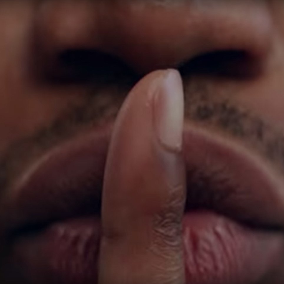 L'image présente un gros plan sur un visage, principalement la zone des lèvres et du nez. Un doigt est placé devant la bouche, formant le geste universel du « chut ». La peau est de teinte foncée, et les lèvres sont bien définies, affichant une légère brillance. Le regard de la personne est probablement dirigé vers l'extérieur de l'image, transmettant une impression de mystère ou de secret. L'arrière-plan est flou, ce qui met l'accent sur le geste et l'expression du visage.