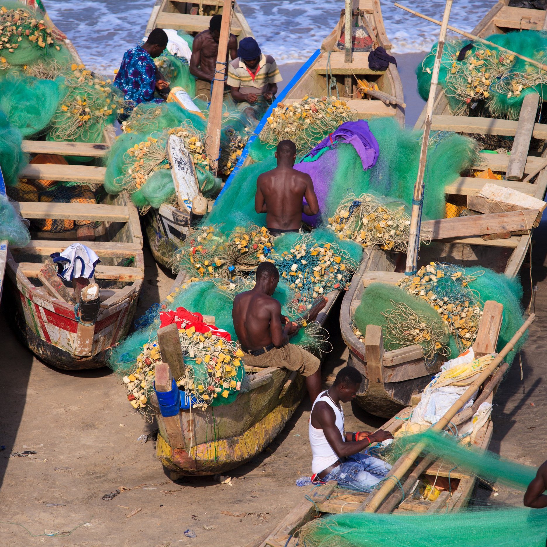 Cette image montre une scène vibrante en bord de mer, où plusieurs pêcheurs sont occupés à travailler sur des bateaux en bois. Les bateaux, peints de différentes couleurs, sont remplis de filets de pêche verts, ainsi que de coquillages et poisson. Les pêcheurs, principalement des hommes, sont torse nu et concentrés sur leurs tâches. L'arrière-plan révèle une mer calme avec des vagues douces, ajoutant une atmosphère de tranquillité à l'activité du port. L'air est imbibé de l'odeur de la mer et du bruit des vagues. Le soleil éclaire la scène, offrant une lumière chaude qui illumine le travail acharné des pêcheurs.