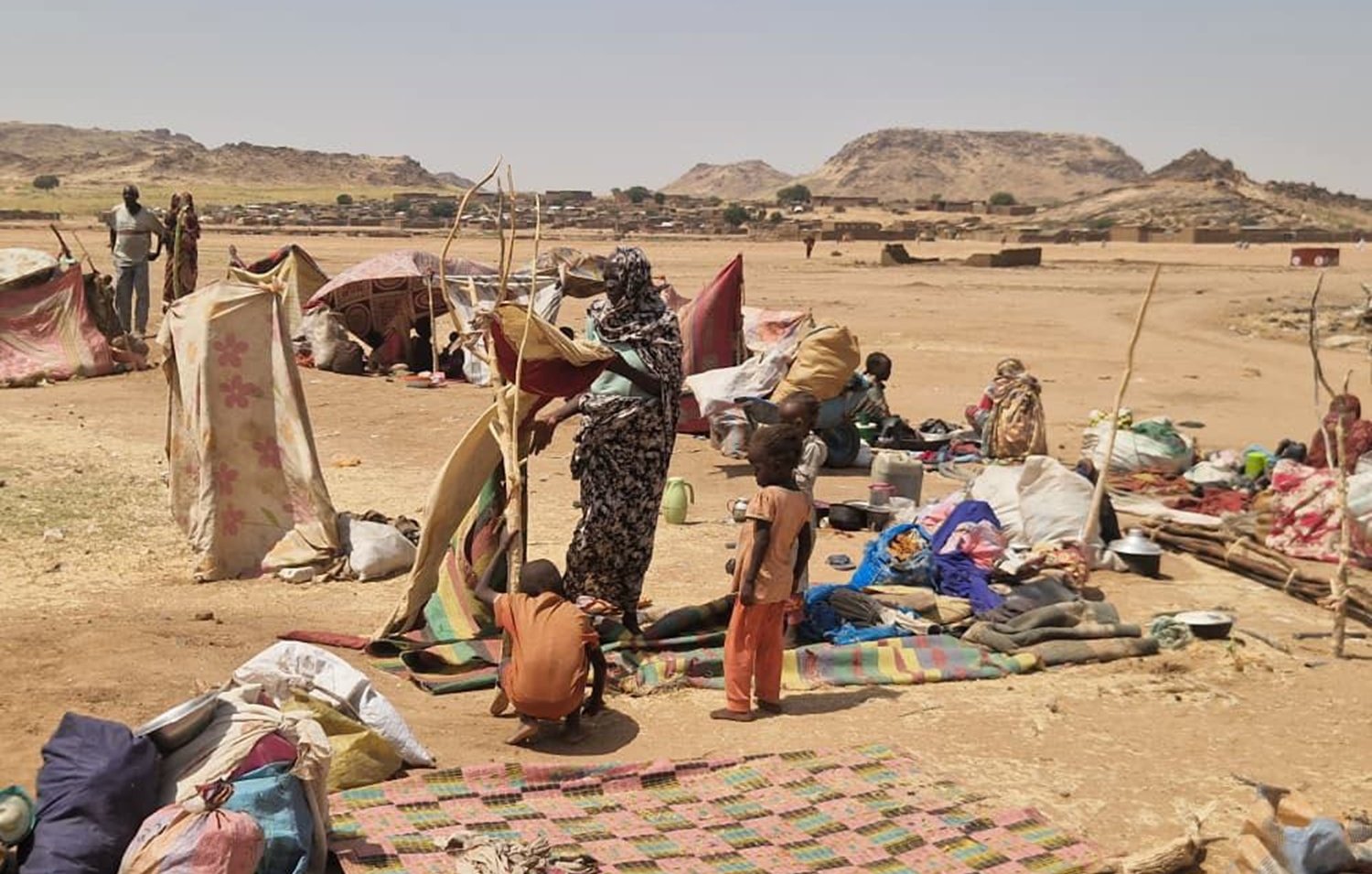 L'image montre un campement dans une région aride et désertique. On peut imaginer une vaste étendue de terrain sec, avec quelques collines en arrière-plan. Des tentes rudimentaires, faites de toile ou de vêtements usagés, sont disposées de manière désordonnée, offrant un semblant de refuge aux personnes qui y vivent. Au sol, des tapis ou des morceaux de tissu sont étalés, tandis que des objets personnels, comme des sacs et des casseroles, sont éparpillés autour. Au centre de la scène, un groupe de personnes est en train de s'activer : une femme semble organiser quelque chose, tandis qu'un enfant debout à côté d'elle observe. L'atmosphère transpire la précarité mais aussi une certaine résilience face aux conditions difficiles. Les couleurs des tissus sont variées, mais plutôt ternes et fanées par le temps. L'ensemble dégage une impression de lutte pour la survie dans un environnement hostile.