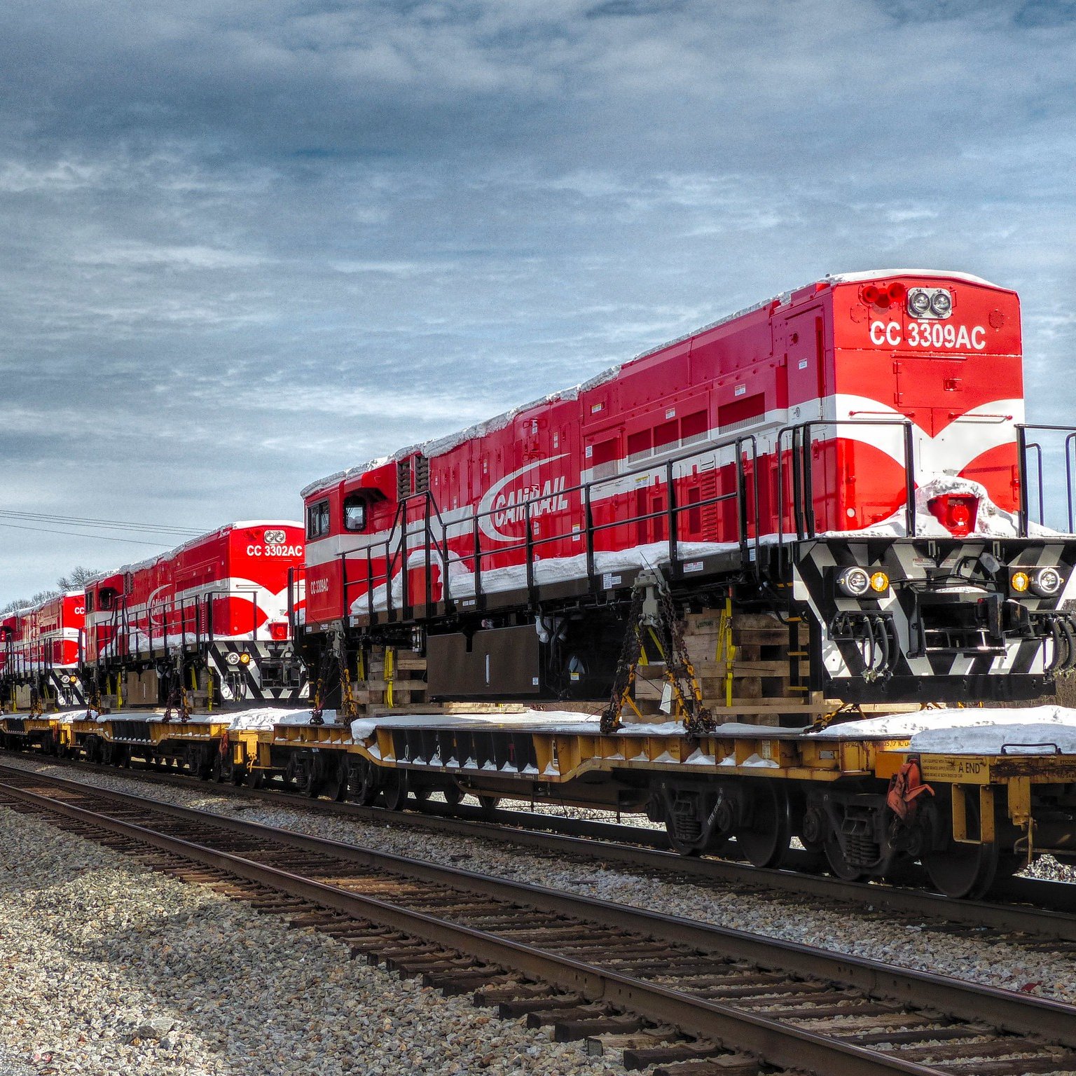L'image montre une série de locomotives de train d'un rouge vif, alignées sur une flatcar (wagon plat) le long des rails. Les locomotives ont un design distinctif avec des motifs blancs et une grande inscription sur le côté. Le ciel est partiellement nuageux, créant un contraste avec la couleur vive des locomotives. Le cadre suggère un environnement industriel, avec des rails de chemin de fer visibles au premier plan et un décor sobre en arrière-plan. L'atmosphère est calme, typique d'une zone où le train est stationné.