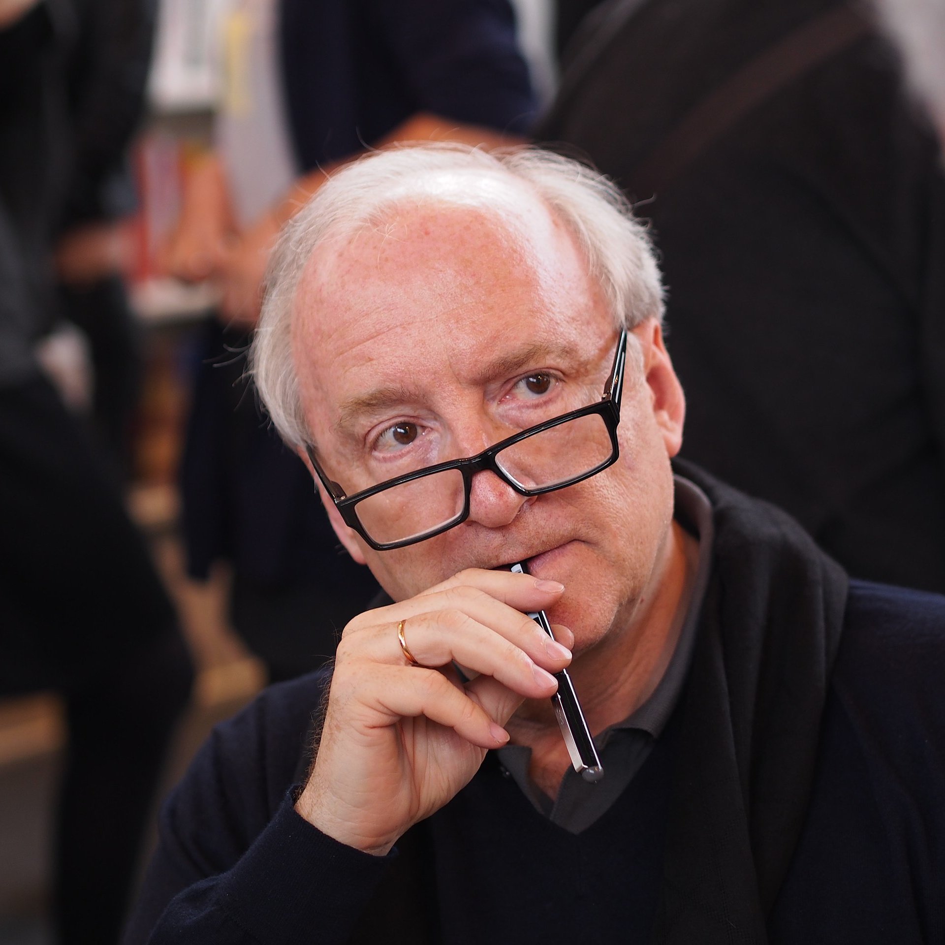 L'image montre un homme assis, vêtu d'un pull sombre, avec des lunettes sur le nez. Il porte une expression pensive, tenant un stylo près de sa bouche, comme s'il était en train de réfléchir ou de considérer quelque chose de manière approfondie. En arrière-plan, on distingue d'autres personnes floues, suggérant une ambiance de discussion ou d'échange, typique d'un événement de rencontre ou de signature de livres. La lumière est douce, créant une atmosphère chaleureuse.