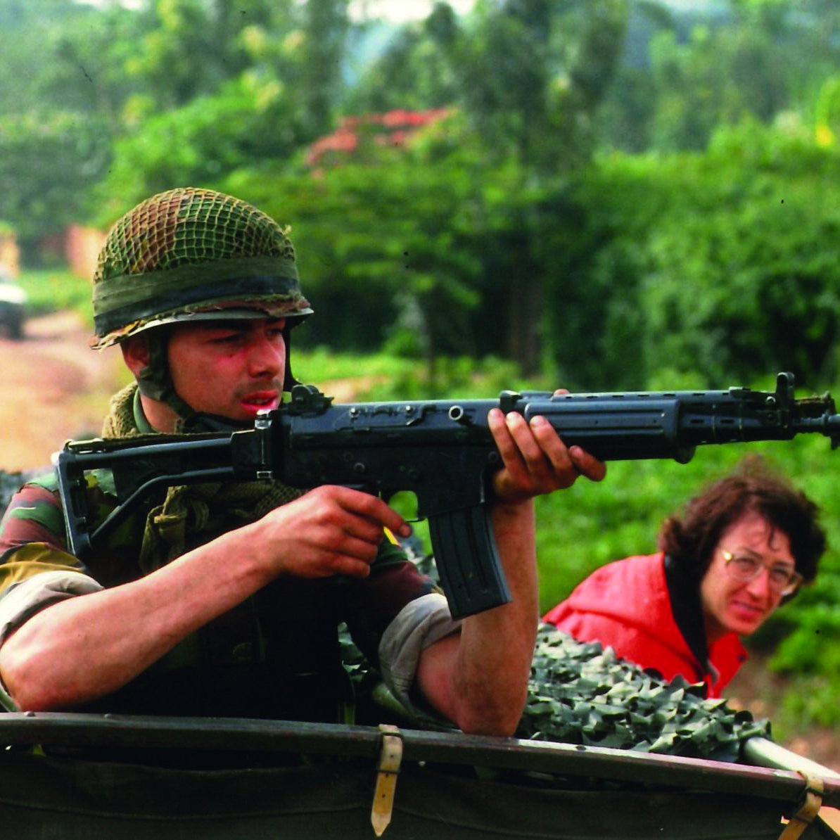 L'image montre un soldat, assis dans un véhicule militaire,tenant un fusil vers l'avant. Il est vêtu d'un uniforme camouflage et porte un casque en treillis. Son visage exprime la concentration et la vigilance, tandis qu'il regarde attentivement son environnement. En arrière-plan, une femme à lunettes observe la scène, semblant préoccupée. La verdure luxuriante entoure le cadre, créant un contraste avec la terre battue sur laquelle se trouve le véhicule. L'atmosphère de l'image est chargée de tension, suggérant un contexte de vigilance et de protection.