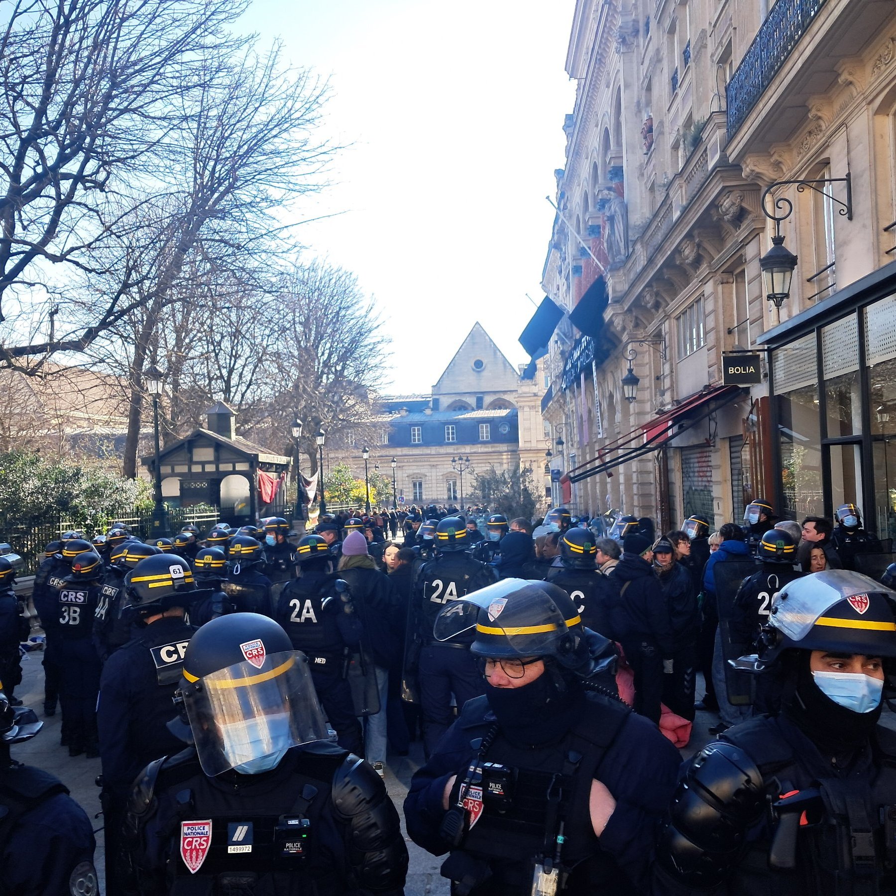 L'image montre une scène urbaine animée. En premier plan, un groupe de policiers en uniforme, équipés de casques et de protections, se tient rassemblé. Leur posture est attentive, suggérant une opération en cours ou une manifestation. À l'arrière-plan, des arbres dénudés et des bâtiments historiques sont visibles, indiquant que la scène se déroule probablement dans une zone urbaine. C'est une journée ensoleillée, avec un ciel clair, apportant une lumière vive sur la scène. L'atmosphère semble tendue, avec des gens qui observent la situation depuis les côtés. On peut percevoir une impression d'encadrement, de sécurité, mais aussi de tension sociale.
