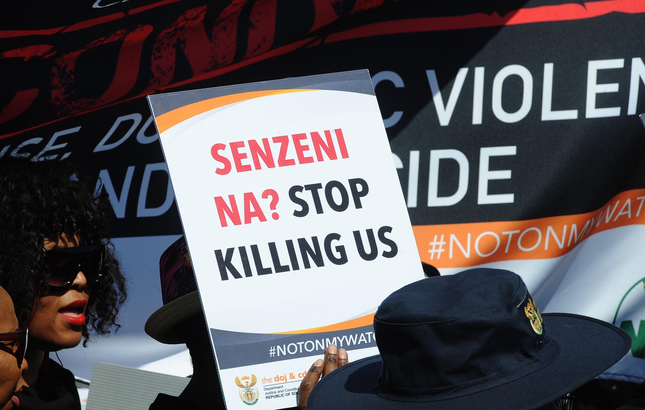 L'image montre un groupe de manifestants rassemblés pour une cause importante. Au premier plan, une personne tient une pancarte avec le slogan « SENZENI NA? STOP KILLING US ». Ce message manifeste un appel urgent à mettre fin à la violence et aux meurtres. En arrière-plan, il y a d'autres bannières et slogans, suggérant une atmosphère de protestation. Les personnes présentes portent des vêtements variés, y compris des chapeaux, et semblent engagées dans la cause qu'elles défendent. L'ambiance générale est celle de la solidarité et de la lutte pour la justice.