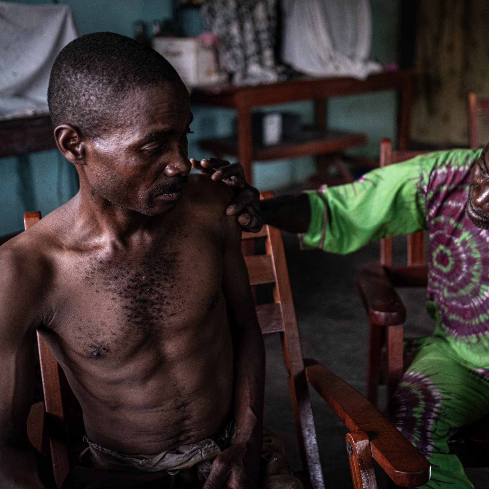 L'image montre deux hommes assis dans des chaises en bois dans une pièce intérieure. L'un des hommes, assis à gauche, a un torse nu et semble pensif. Son expression montre une certaine vulnérabilité. L'autre homme, à droite, est vêtu d'une robe de couleur verte et violette, et il semble s'adresser au premier avec attention, posant une main réconfortante sur son épaule. La lumière dans la pièce est douce, créant une atmosphère intimiste. En arrière-plan, on peut voir des objets flous disposés sur des tables, ce qui suggère un environnement familier et intime.