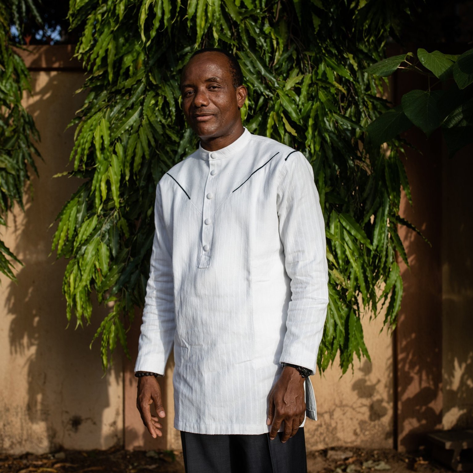 L'image montre un homme debout devant un arrière-plan de feuillage vert dense. Il porte une tunique blanche à col montant et un pantalon sombre. Son sourire dégage une attitude amicale et confiante. Les arbres autour de lui ajoutent une ambiance naturelle, avec des branches feuillues qui encadrent sa silhouette. Le fond est constitué de murs dans des tons nude, apportant une touche chaleureuse à la scène. La lumière naturelle semble douce, créant une atmosphère sereine.