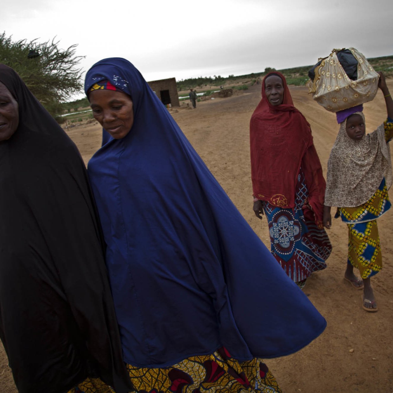 L'image représente un groupe de cinq femmes marchant sur un chemin de terre. Elles portent des vêtements traditionnels colorés, avec des motifs variés. Certaines femmes ont un voile couvrant leur tête, et une jeune fille à l'arrière transporte un objet sur sa tête, probablement un panier. Le paysage environnant est sec et désertique, avec quelques arbustes épars en arrière-plan, et le ciel est couvert, suggérant une ambiance calme et sereine. L'ensemble transmet une impression de solidarité et de culture, avec un accent sur les traditions vestimentaires.