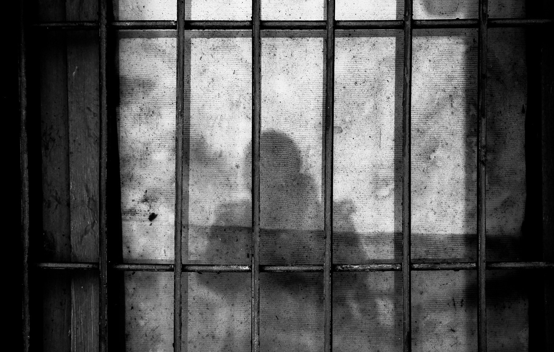 L'image présente une fenêtre avec des barreaux, offrant une vue obscure et mystérieuse. À travers le verre sale et légèrement opaque, une silhouette se découpe, créant une ambiance intrigante. L'éclairage est doux, se déversant à travers la saleté du verre, ce qui donne une atmosphère presque nostalgique. Les contours de la silhouette apparaissent flous, laissant place à l'imagination sur ce qui se trouve derrière cette barrière. Les textures des barreaux et du verre ajoutent une dimension tactile à l'ensemble.