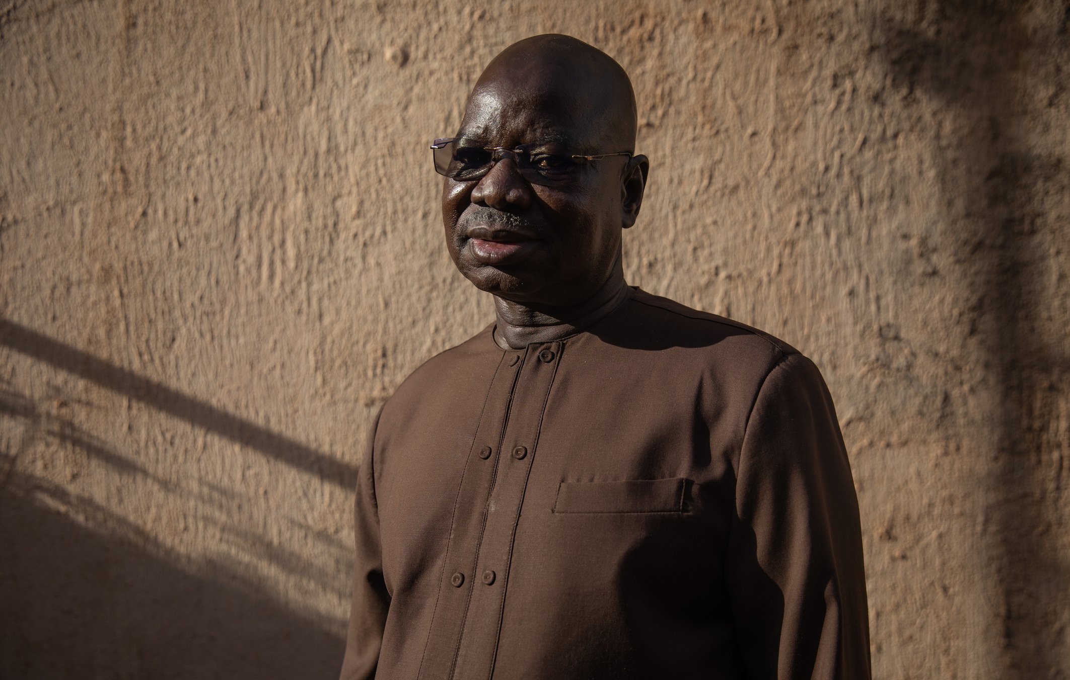 L'image présente un homme d'âge mûr, avec un teint foncé et des lunettes. Il porte une tenue sombre, légèrement scintillante, adaptée à un environnement formel ou culturel. Son expression est sereine et réfléchie. Le fond est constitué d'un mur en terre ou en adobe, éclairé par une lumière naturelle douce, créant une ambiance chaleureuse. Des ombres longues suggèrent qu'il est en fin de journée, ajoutant à la profondeur de l'image.