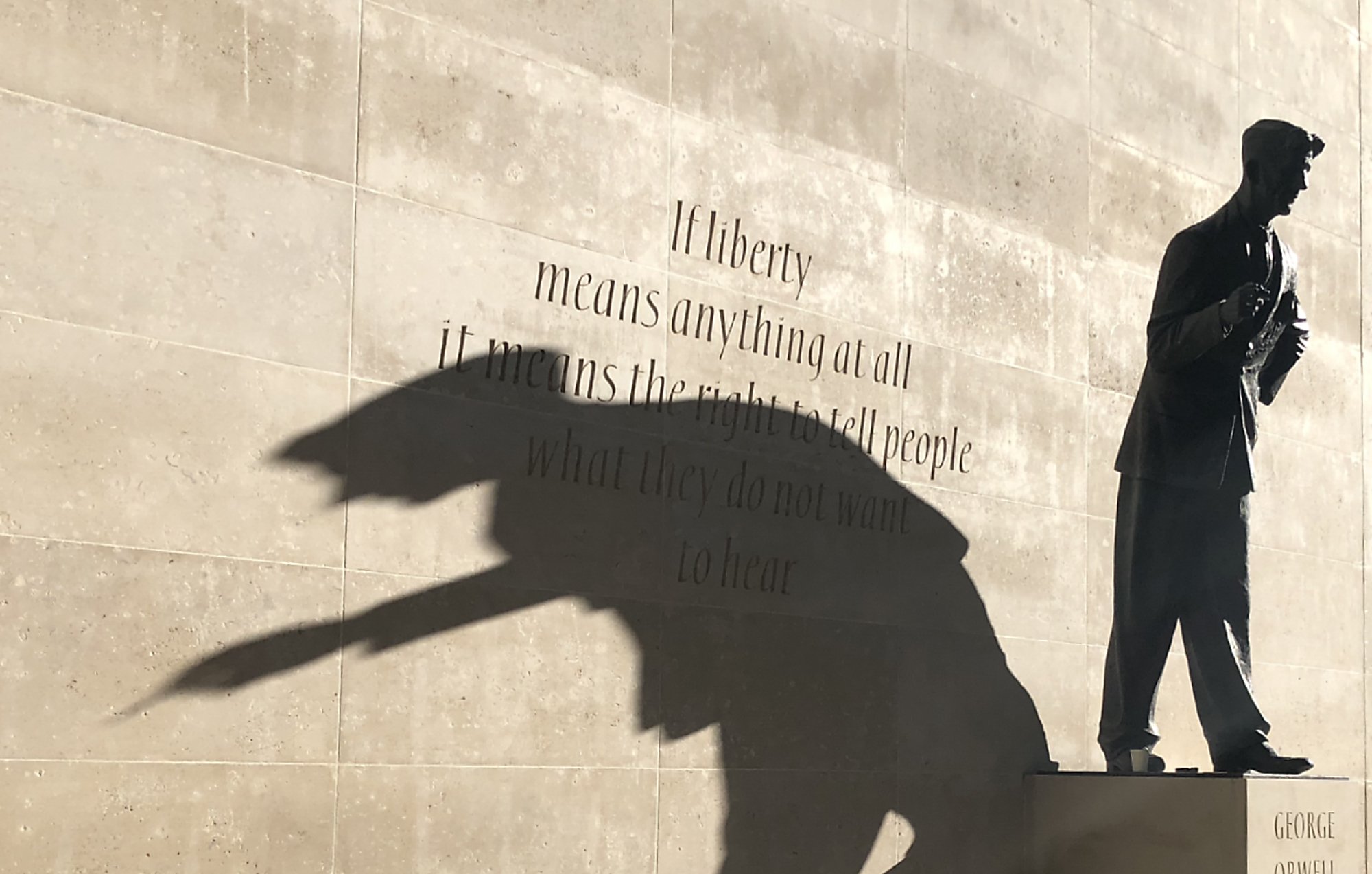 L'image représente une statue d'un homme debout, probablement un auteur ou un penseur, sur un socle. La statue est en bronze, et son ombre projette une forme dynamique sur le mur derrière elle. Ce mur est en pierre lisse, et il porte une citation célèbre qui commence par « If liberty means anything at all... », reflétant des thèmes de liberté et d'expression. L'ensemble crée une atmosphère réfléchie et inspirante, incitant à considérer la profondeur des mots inscrits ainsi que le rôle de la statue.