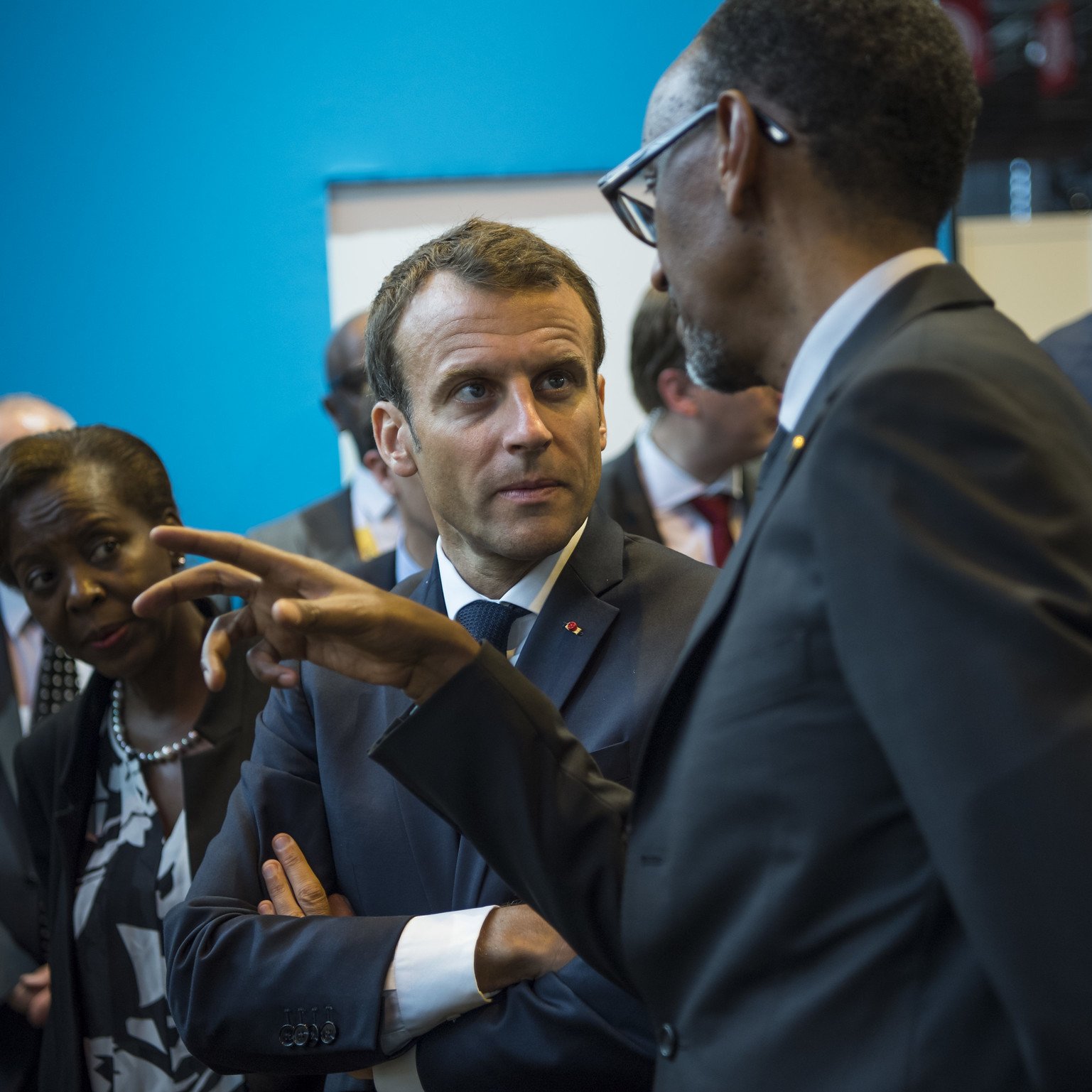L'image montre un groupe de personnes en conversation dans un environnement qui semble formel. Au premier plan, un homme en costume sombre, probablement un leader, discute avec un autre homme, qui porte également un manteau formel. Le premier homme a une expression attentive, se concentrant sur la discussion, tandis que le second gesticule avec sa main, comme s'il faisait un point important. En arrière-plan, une femme les observe, affichant une expression de sérieux. Le décor est composé de murs d'un bleu vif, ce qui crée un contraste avec les costumes sombres des personnages. L'atmosphère semble professionnelle et engagée, suggérant une rencontre importante.