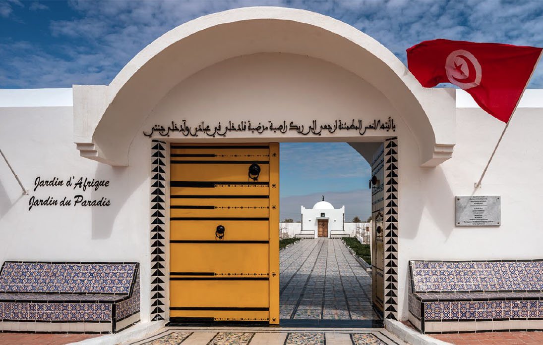 Imaginez un lieu paisible et accueillant, avec une grande porte jaune ornée de motifs géométriques. Cette porte s'ouvre sur un jardin magnifique, symbolisant la beauté et la diversité de la nature. Deux bancs placés de chaque côté de l'entrée, recouverts de carrelage coloré, invitent à la détente. Au-dessus de la porte, une inscription en arabe et en français nous invite à découvrir ce jardin, intitulé "Jardin d'Afrique, Jardin du Paradis". De chaque côté, un drapeau flotte, ajoutant une touche de couleur vive à la scène. Le ciel est lumineux et parsemé de nuages, apportant une lumière douce qui souligne la sérénité de cet endroit. L'ambiance générale est calme et harmonieuse, évoquant un espace de ressourcement et de contemplation.
