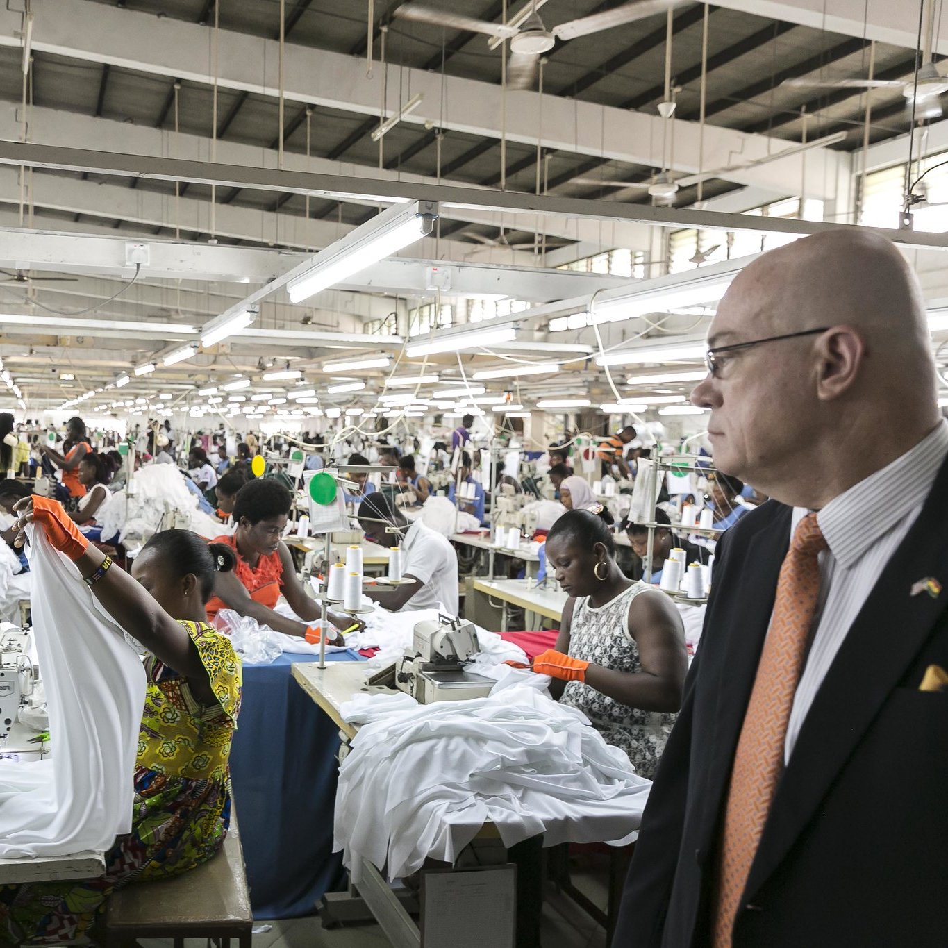 L'image montre une grande usine de couture animée. De nombreux ouvriers, principalement des femmes, sont occupés à coudre des vêtements, en particulier des chemises blanches. Ils travaillent en équipe autour de tables de couture, utilisant des machines à coudre. L'ambiance est dynamique, avec des mouvements rapides des mains et des tissus qui flottent. À droite, un homme en costume observe la scène, se tenant debout et faisant face au travail en cours. L'usine est bien éclairée, avec des lumières suspendues au plafond, et les murs sont de couleur claire, créant un espace de travail ouvert et organisé.
