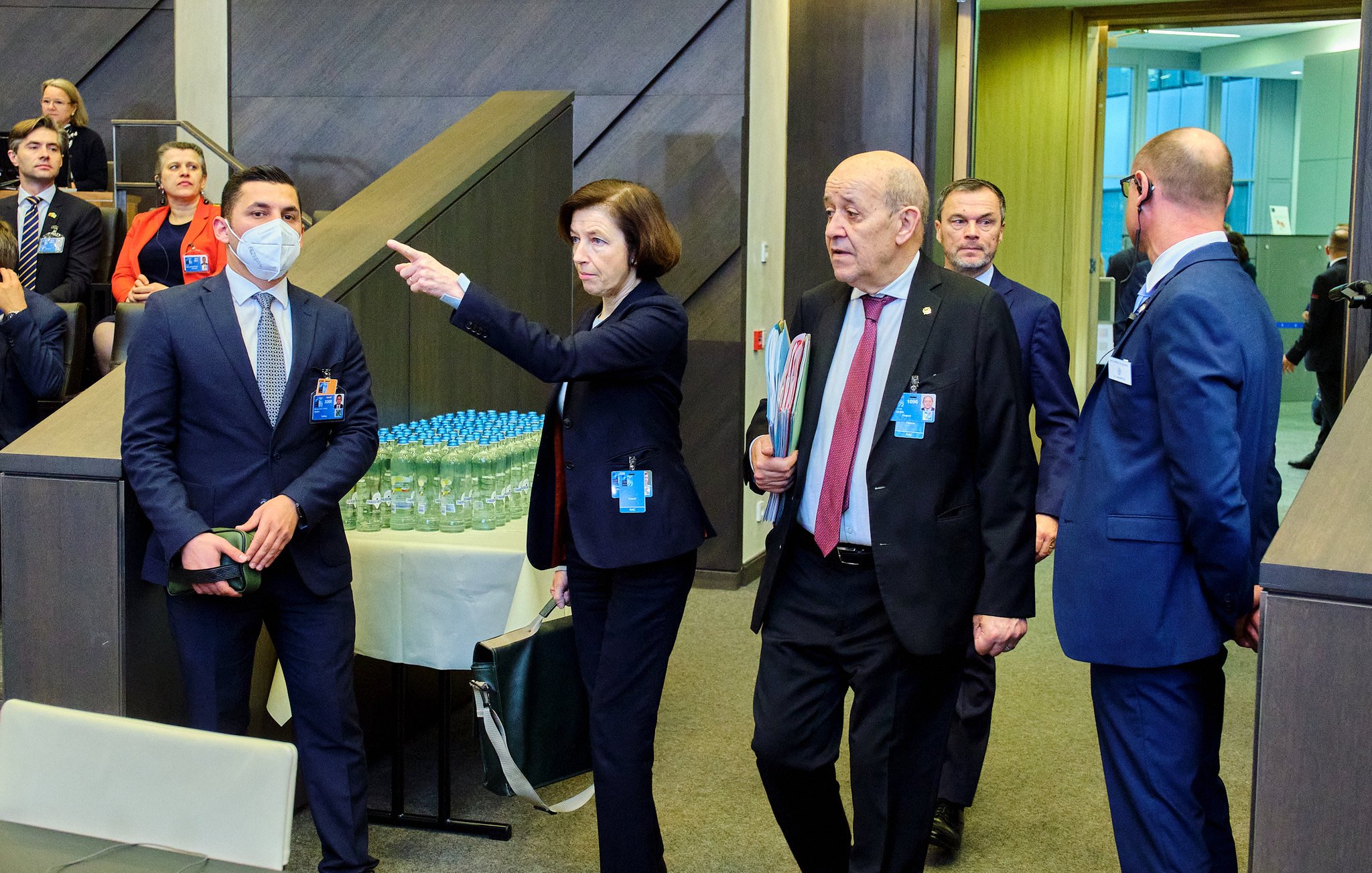 Dans cette scène, un groupe de personnes se déplace à l'intérieur d'un bâtiment moderne, probablement lors d'une réunion ou d'un événement officiel. Au premier plan, une femme, vêtue d'un costume professionnel et portant un document à la main, pointe dans une direction, semblant donner des indications. À ses côtés, un homme portant un masque de protection écoute attentivement, tandis qu'un autre homme, en costume noir, marche à ses côtés, apparemment engagé dans une conversation. En arrière-plan, d'autres participants sont visibles, suggérant une atmosphère animée et collaborative. Des bouteilles d'eau sont disposées sur une table à proximité, signalant la durée de l'événement. Le décor est contemporain, avec des murs en bois et des éléments de design épuré.