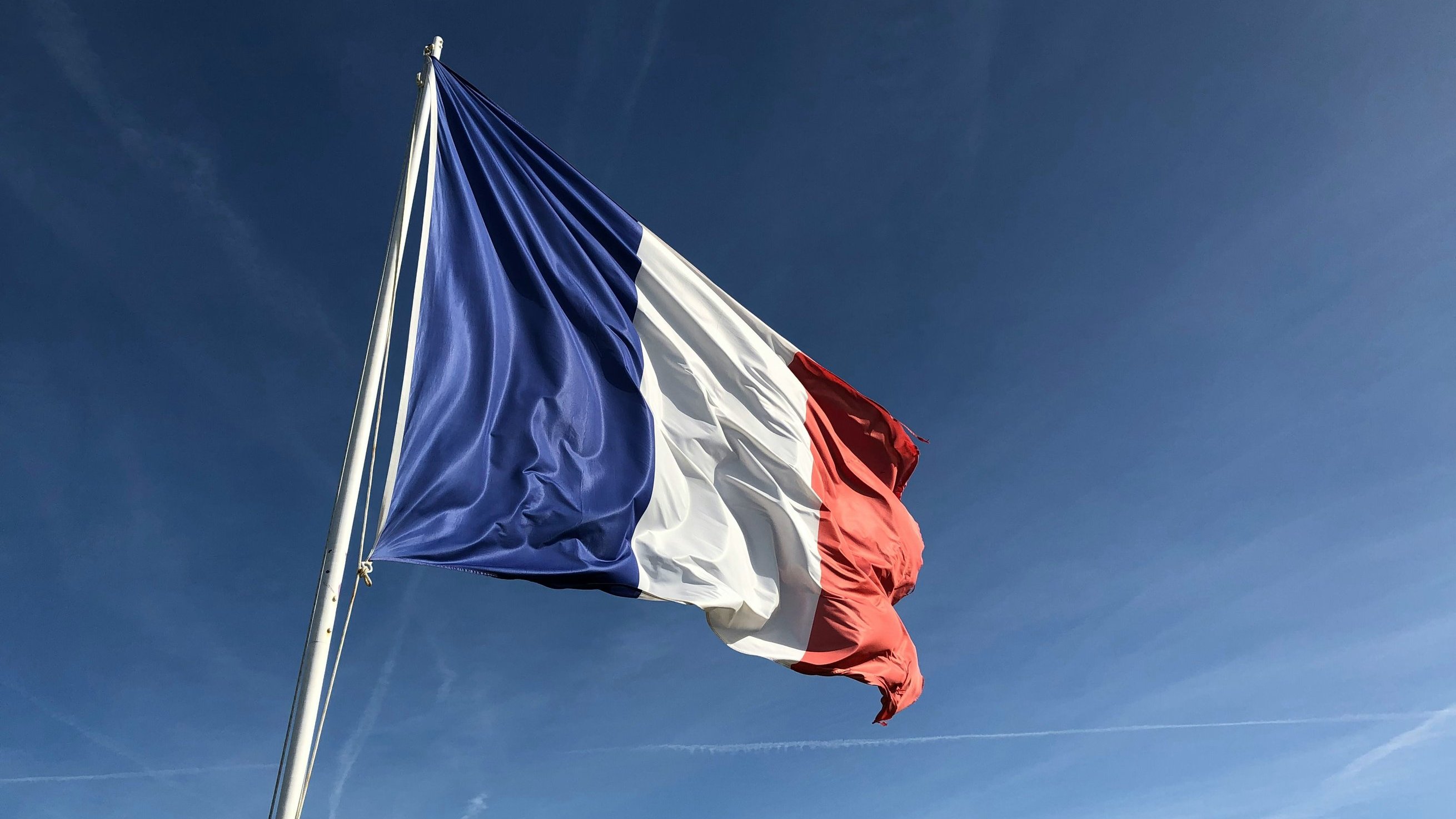 L'image montre un drapeau, qui est le drapeau tricolore français. Il est composé de trois bandes verticales : bleu à gauche, blanc au centre et rouge à droite. Le drapeau est agité par le vent, ce qui donne une impression de mouvement. À l'arrière-plan, on peut imaginer un ciel clair, probablement d'une belle journée ensoleillée, avec quelques traînées de nuages dans un bleu vif. Le contraste entre les couleurs du drapeau et la luminosité du ciel crée une image frappante et symbolique.