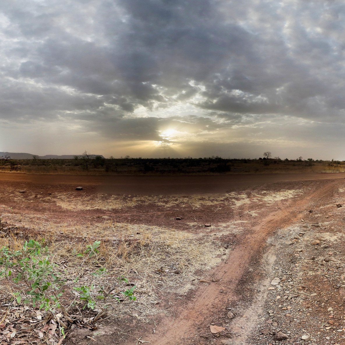Imaginez un vaste paysage ouvert, dominé par des tons chauds de terre et des nuances de beige. Le sol est sec et parsemé de petites pierres, avec des herbes jaunies qui émergent de la terre aride. À l'horizon, un ciel nuageux s'étend, teinté de gris avec des rayons de lumière qui percent, annonçant un coucher de soleil. La lumière crépusculaire crée une ambiance paisible, tandis que la nature environnante semble calme et tranquille. Les silhouettes lointaines de collines ajoutent une dimension au paysage, renforçant le sentiment d'immensité et d'isolation.