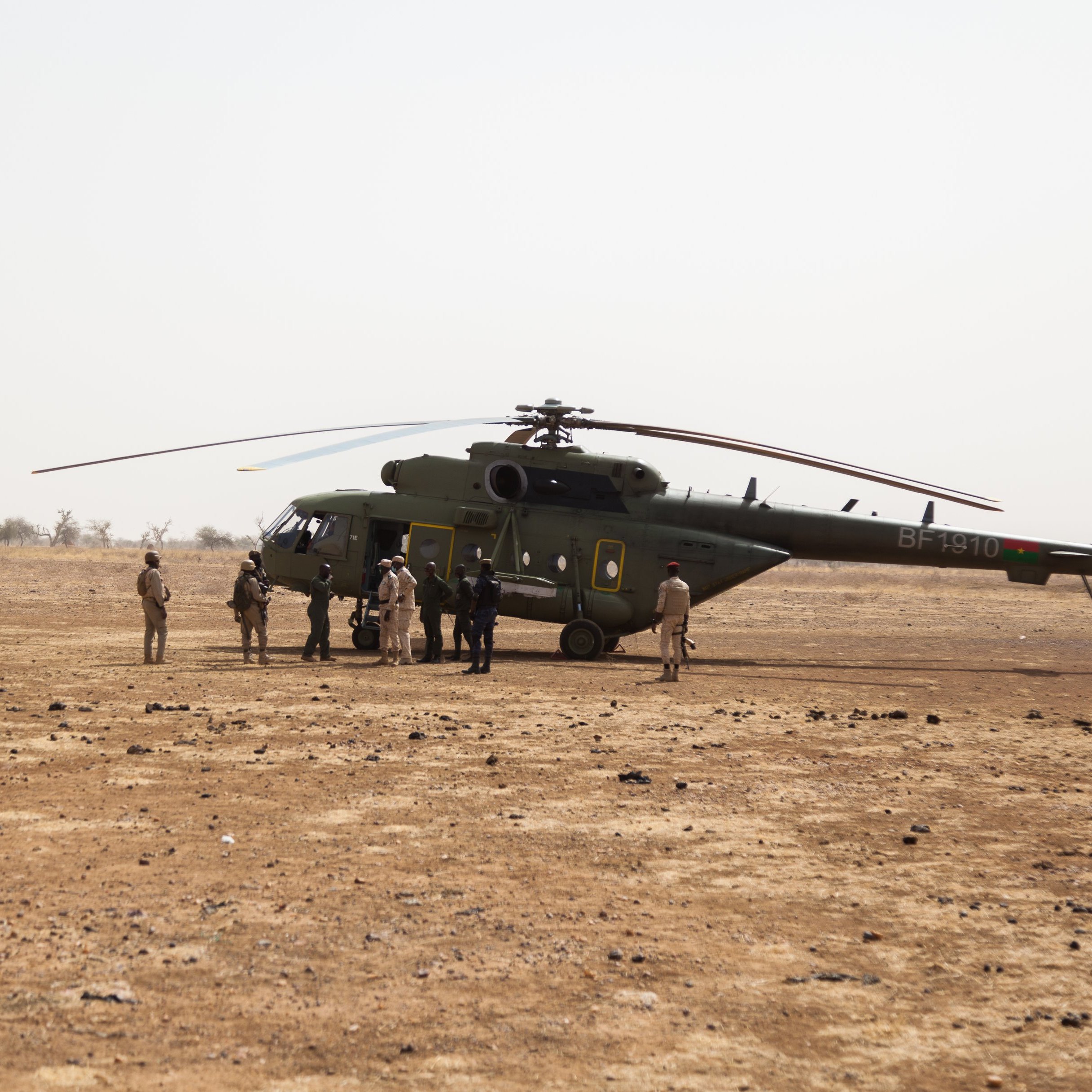 L'image montre un hélicoptère posé sur un terrain désertique, avec un sol caillouteux et sec. Autour de l'hélicoptère, plusieurs personnes sont rassemblées, certaines en uniforme militaire. La scène semble se dérouler dans une région aride, avec peu de végétation en arrière-plan.