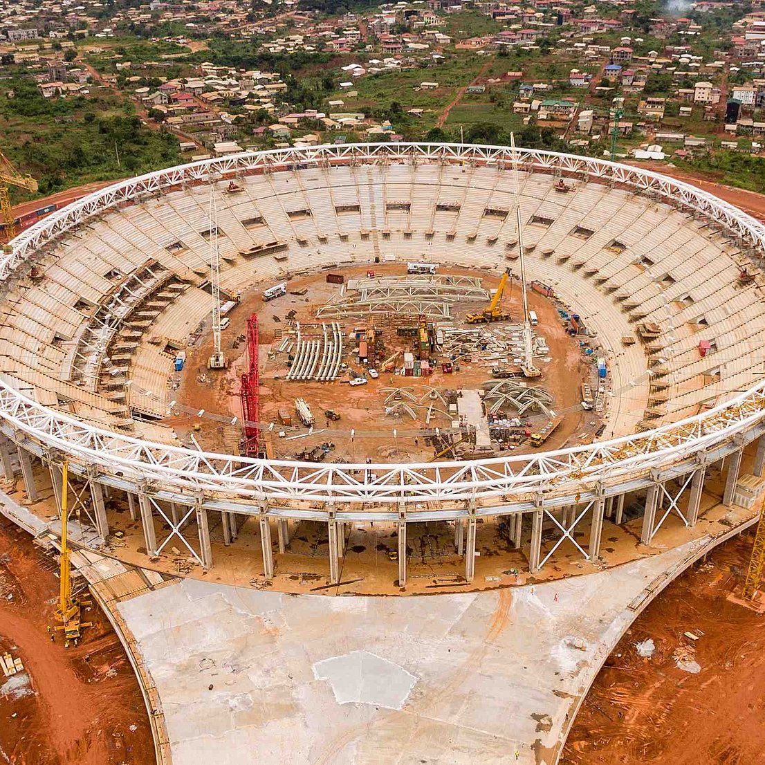 L'image montre un stade en cours de construction, vu d'en haut. Le stade a une forme ovale et un grand espace intérieur qui est encore vide, avec des structures en cours d'installation. Autour du stade, on peut voir un terrain rouge, probablement une terre fouillée pour la construction. À l'extérieur de la structure, il y a des machines de construction et des matériaux éparpillés. À l'arrière-plan, des maisons et des collines sont visibles, ce qui donne une idée de l'environnement urbain et naturel qui entoure le stade. L'ensemble de la scène évoque une ambition de développement et de modernisation.