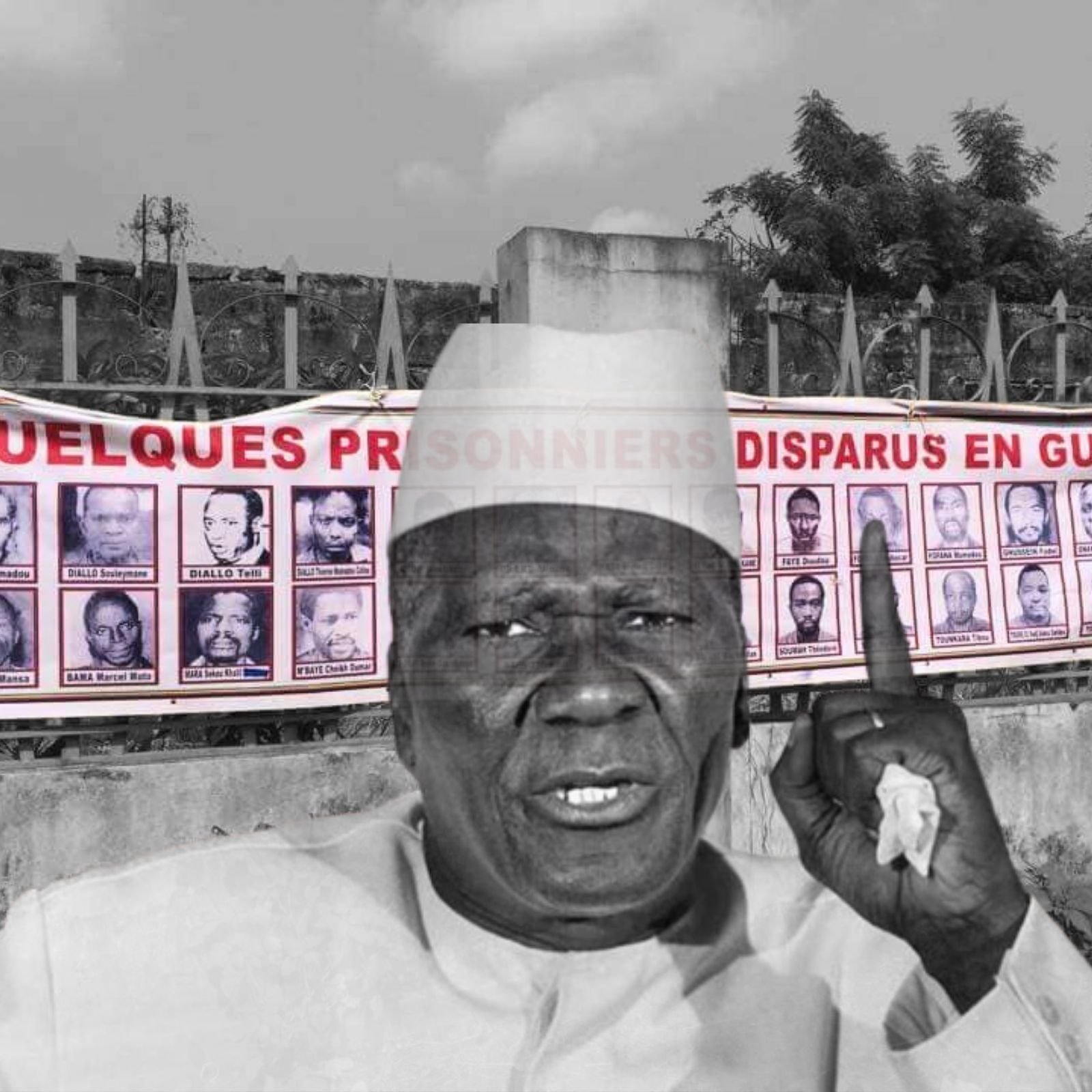 L'image montre un homme d'âge mûr, vêtu d'une tenue traditionnelle et portant un chapeau. Il lève un doigt en signe d'alerte ou de demande d'attention. En arrière-plan, on peut voir une grande banderole accrochée sur un mur. Cette banderole affiche des portraits de plusieurs personnes, accompagnés d'un texte qui fait référence à des prisonniers disparus en Guinée. L'ambiance de l'image évoque un appel à ne pas oublier ces disparitions et à sensibiliser le public à cette problématique.