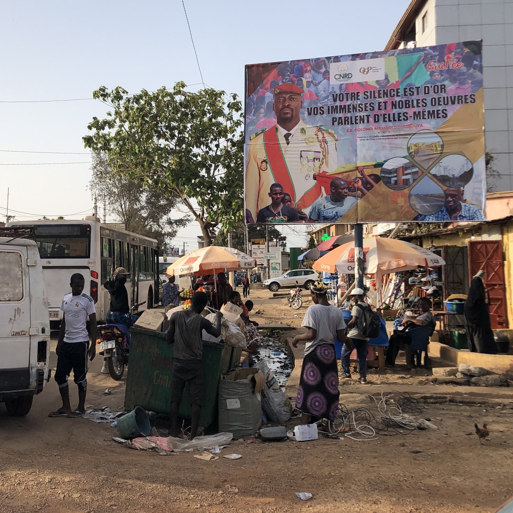 L'image représente une scène de rue animée dans une ville. Au premier plan, on voit une camionnette blanche à l'arrière, avec des détails usés et des portes ouvertes. À gauche, un bus de transport en commun est stationné. Plusieurs personnes se déplacent sur la chaussée, certaines vendant des marchandises sous des parasols colorés. Il y a également des bacs de récupération et divers déchets sur le sol, indiquant une ambiance de marché. En arrière-plan, un grand panneau publicitaire présente des visages de personnes et un message en français, affichant un slogan qui semble inciter à l'action ou au changement. Les arbres et les fils électriques ajoutent à l'atmosphère urbaine du lieu.