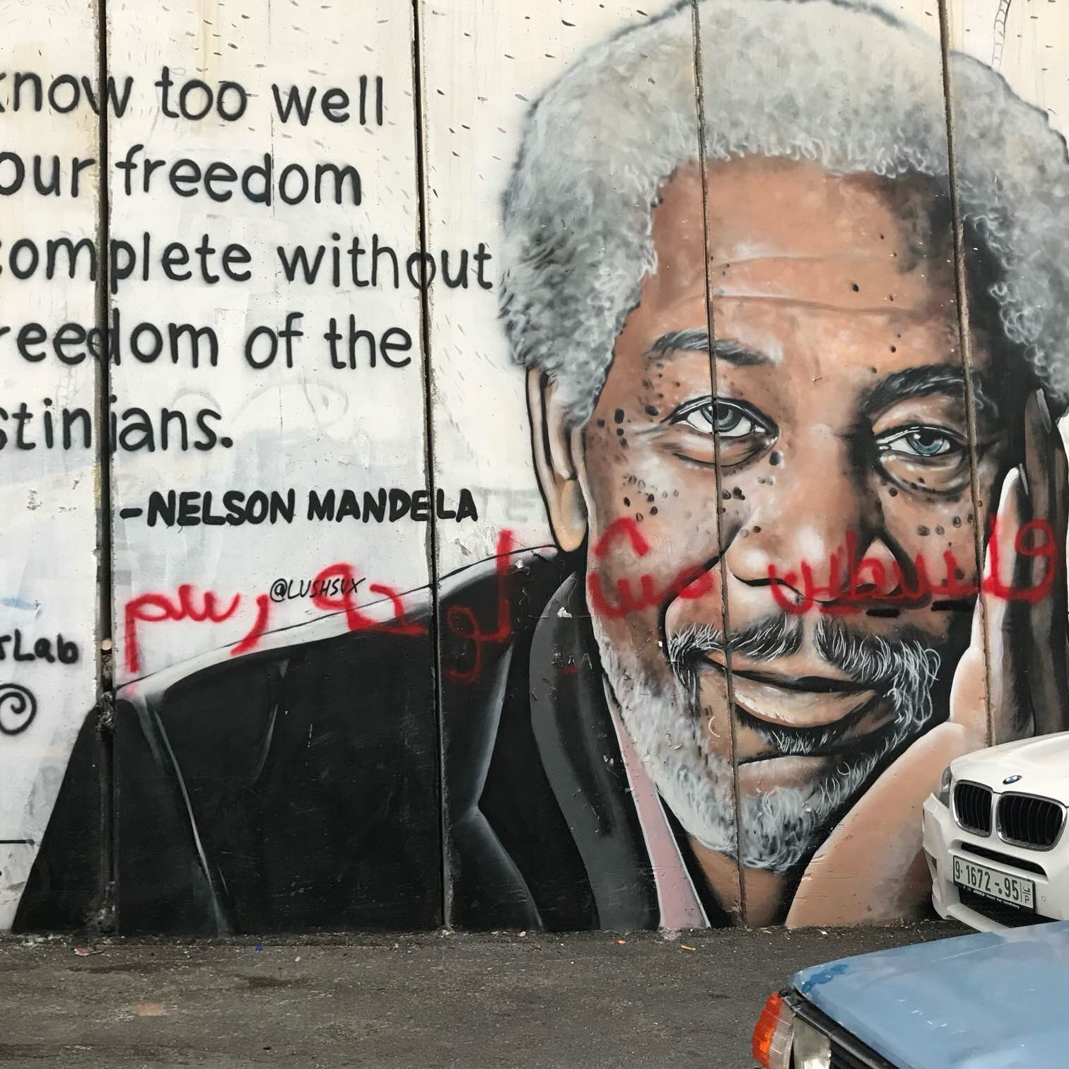 L'image représente un mur sur lequel est peint un mural poignant. À gauche, on voit un portrait détaillé d'un homme âgé avec des cheveux grisonnants et une expression réfléchie. À droite du portrait, une citation attribuée à Nelson Mandela évoque l'importance de la liberté des Palestiniens pour garantir notre propre liberté. Le texte est en anglais, avec un style graphique qui attire l'attention. En bas, quelques graffitis ajoutent une touche de couleur et de dynamisme à l'ensemble. L'ambiance est à la fois engageante et réfléchie, signifiant une connexion profonde entre les luttes pour la liberté.