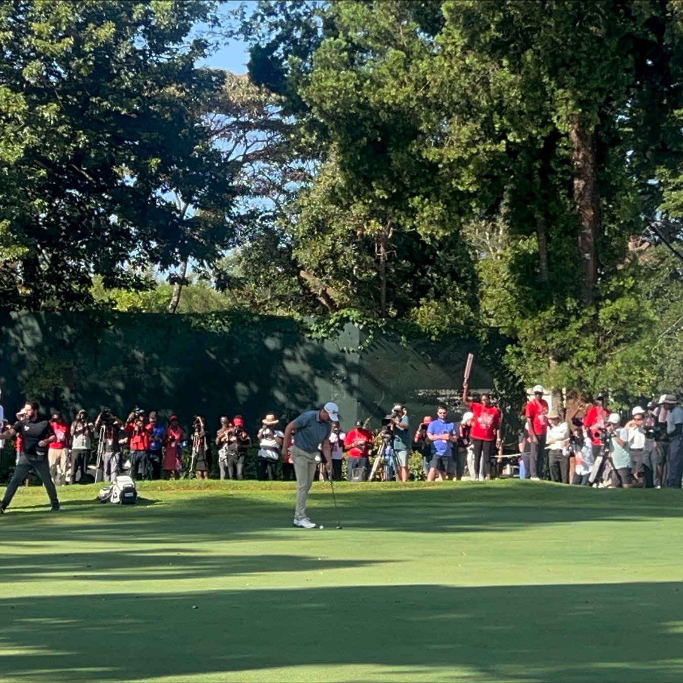 Imaginez un vaste terrain de golf, baigné par la lumière du soleil. Au premier plan, deux joueurs se concentrent sur le putting green, faisant preuve de délicatesse et de précision. L'un d'eux est en position de putt, tandis que l'autre observe attentivement. À l'arrière-plan, des spectateurs en tenue décontractée, vêtus de couleurs variées, attendent avec excitation, certains filmant ou prenant des photos. Autour du green, de grands arbres fournissent de l'ombre, ajoutant à la beauté de l'environnement. À droite, on aperçoit un bâtiment avec des balcons, où d'autres personnes sont rassemblées pour profiter du spectacle. L'atmosphère est vivante, remplie de murmures, de rires et de l'anticipation d'un moment décisif dans le jeu.