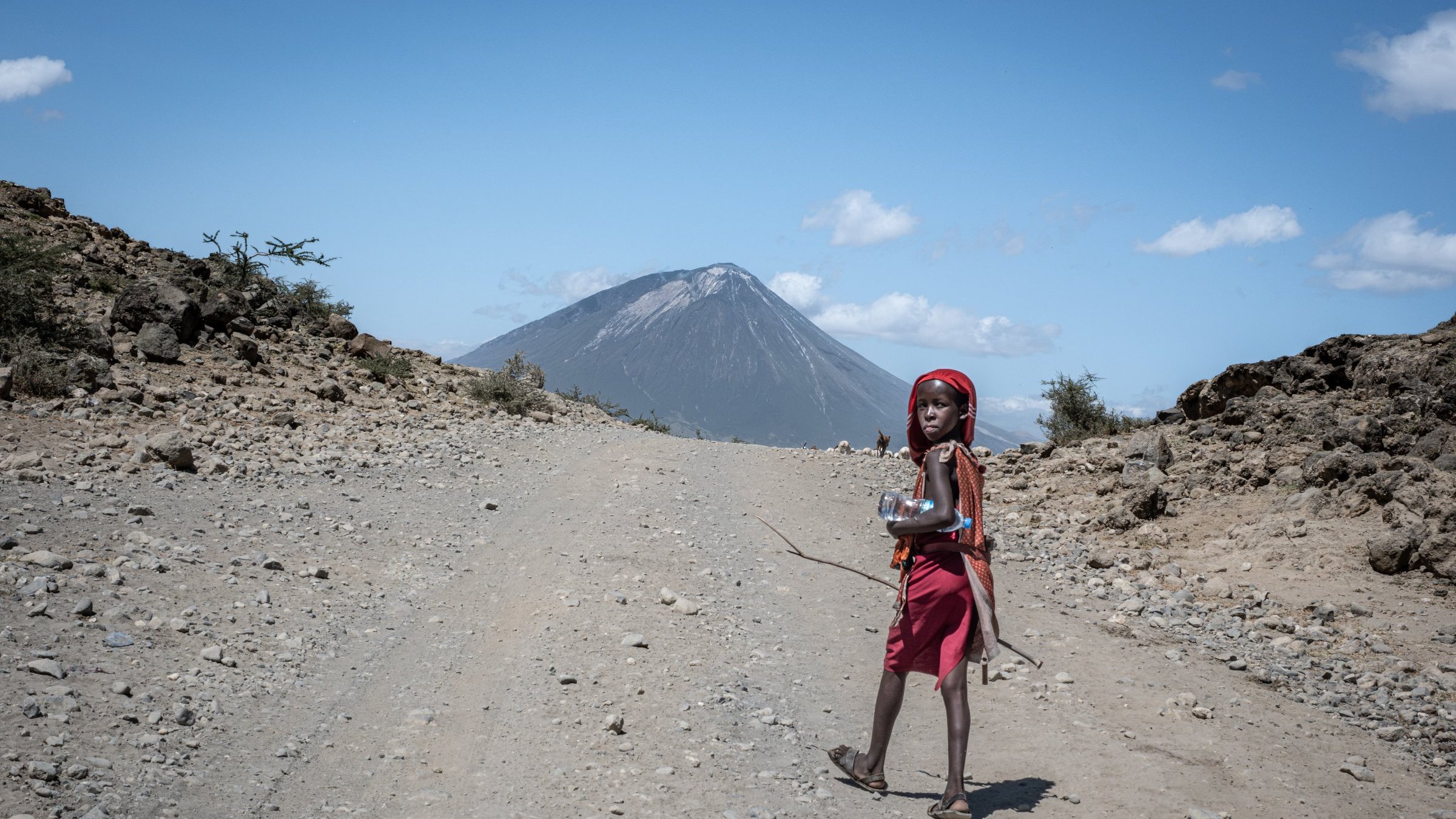 L'image montre un enfant marchant sur un chemin de terre. Il porte une robe rouge et un foulard, et il semble porter un petit récipient ou une bouteille dans ses mains. En arrière-plan, on aperçoit un volcan majestueux, dont le sommet est légèrement nuageux sous un ciel bleu clair parsemé de quelques nuages blancs. Le terrain autour est rocailleux et aride, avec quelques petits buissons éparpillés. L'ensemble de la scène dégage une atmosphère de nature sauvage et éloignée.