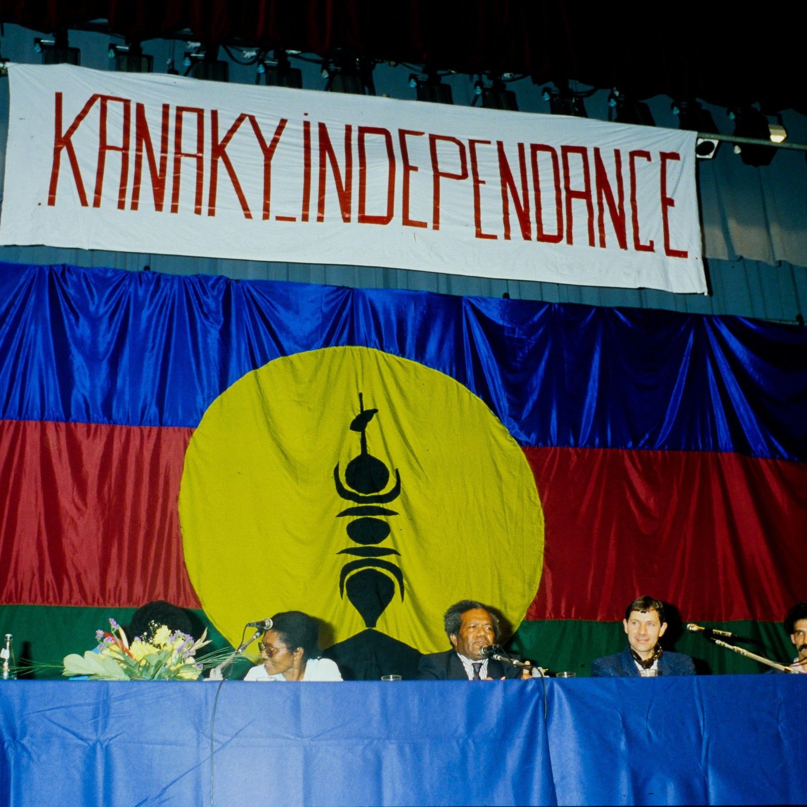 L'image présente une scène de réunion ou de conférence, où des personnes sont assises derrière une table. En arrière-plan, il y a un grand drapeau avec des couleurs vives : un rouge et un bleu, et un cercle jaune au centre. Dans ce cercle, on peut distinguer un symbole noir, précisant ainsi l'identité culturelle. Au-dessus de la scène, une banderole arbore le mot "KANAKY INDEPENDANCE" écrit en lettres rouges, exprimant un message fort lié à l'indépendance. Les participants semblent engagés et sont en discussion, créant une atmosphère de détermination et de solidarité.
