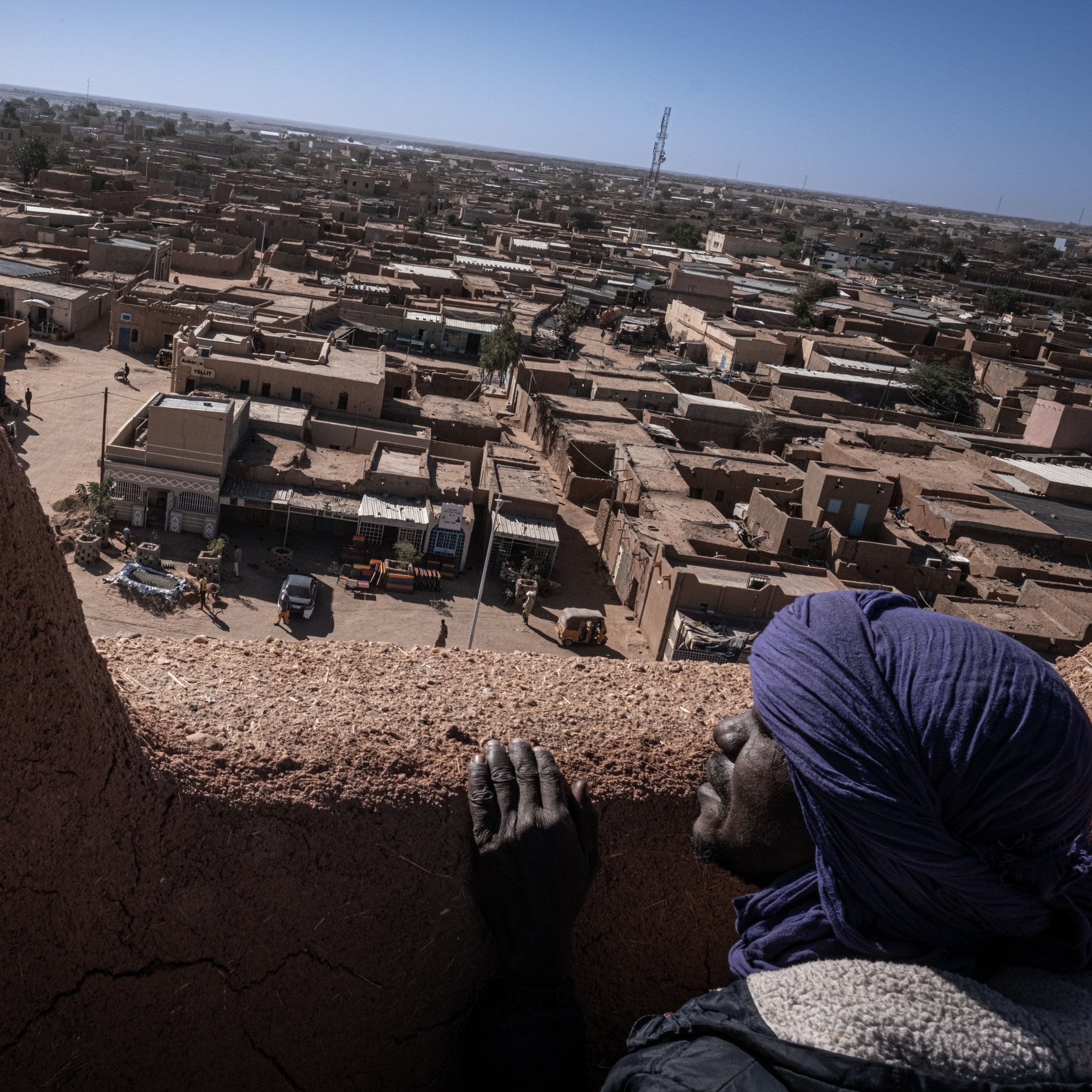 L'image présente une vue panoramique d'une ville saharienne. Au premier plan, une personne au visage marqué par le temps s'appuie sur un mur en terre. Elle porte un turban et semble contempler le paysage. En arrière-plan, des maisons en adobe s'étendent à perte de vue, avec des toits plats et des teintes ocres. On peut apercevoir des petites routes et des véhicules qui circulent. L'horizon est dégagé, avec quelques structures modernes qui contrastent avec l'architecture traditionnelle. L'atmosphère évoque à la fois la chaleur du désert et la tranquillité d'une ville endormie.