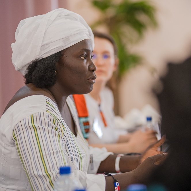L'image montre une femme assise à une table lors d'une réunion ou d'une discussion. Elle porte une tenue traditionnelle, avec une chemise à rayures et un foulard blanc sur la tête. Son expression est sérieuse et concentrée, et elle semble engagée dans la conversation. À côté d'elle, une autre femme est présente, mais elle est floue en arrière-plan. On peut apercevoir des bouteilles d'eau sur la table, ainsi qu'une ambiance chaleureuse et collaborative. Des éléments de verdure, comme une plante, ajoutent une touche de couleur à la scène.