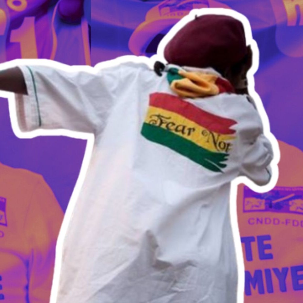 The image features a figure wearing a white shirt with vibrant colors and a design on the back. The background includes repeated images of a man, possibly a political figure, smiling and wearing a hat with a logo. The overall color scheme is purple, enhancing a dynamic atmosphere. The shirt's imagery suggests a message of confidence or defiance.