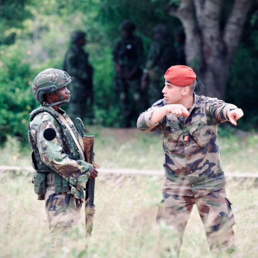 L'image montre deux militaires en discussion dans un environnement extérieur. À gauche, un soldat vêtu d'un uniforme camouflage, portant un casque et tenant un fusil. À droite, un autre militaire, qui porte une tenue camouflage et un béret rouge. Il semble gesticuler en indiquant une direction. En arrière-plan, on aperçoit d'autres soldats flous, ce qui suggère qu'ils sont dans une zone d'entraînement ou opérationnelle, entourée de végétation et d'arbres. L'ambiance est sérieuse et dénote une situation de coordination ou de briefing entre les membres de l'armée.