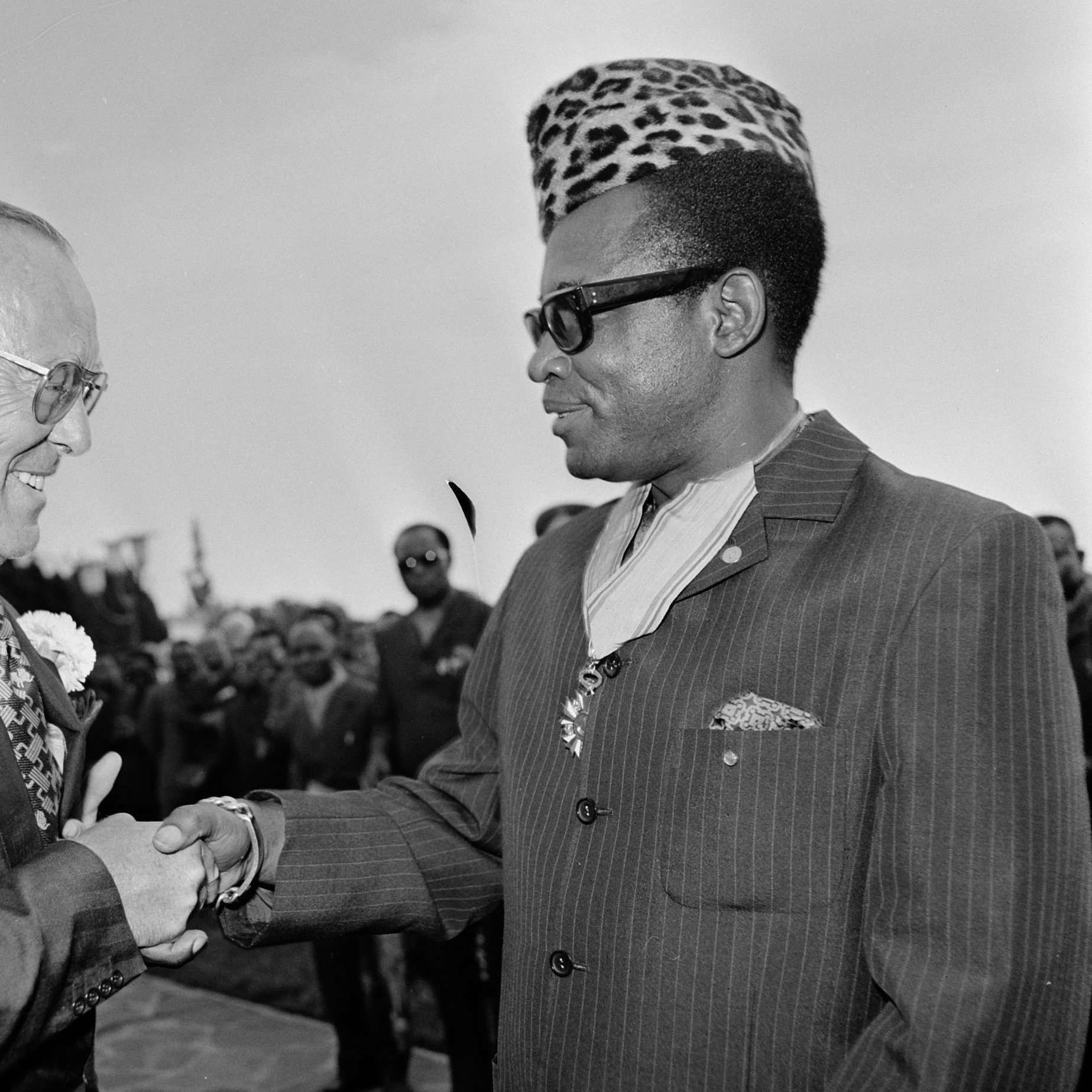 L'image montre une scène de rencontre entre deux hommes souriants, se serrant la main dans un cadre formel et convivial. Le premier homme porte un costume sombre avec une chemise à motifs fleuris et des lunettes, tandis que le second, qui semble être une figure importante, arbore un élégant costume rayé et un chapeau à motifs distinctifs, évoquant une riche culture. En arrière-plan, un groupe de personnes observe la scène, ajoutant à l'impression d'un événement important. L'atmosphère est chaleureuse et amicale, suggérant une rencontre diplomatique ou un événement marquant.