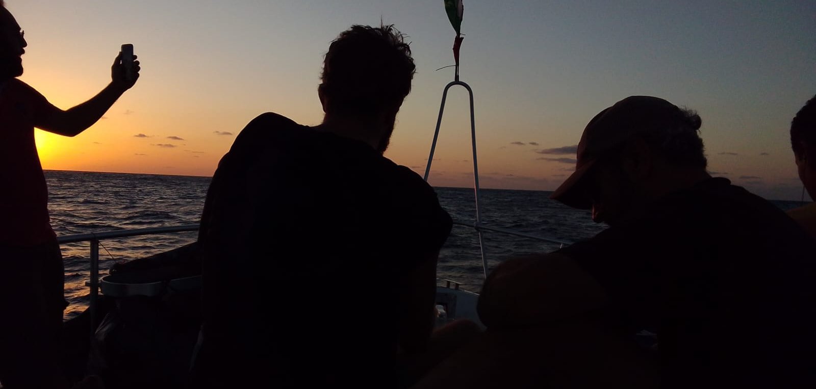 The image shows a group of people on a boat at sunset. The sky is painted in warm hues of orange and yellow as the sun sets on the horizon. Two people are visible, sitting with their backs to the camera, seemingly enjoying the view. One person is standing and holding a phone, possibly taking a photo. The water appears calm, reflecting the colors of the sky. The overall atmosphere is serene and relaxed.